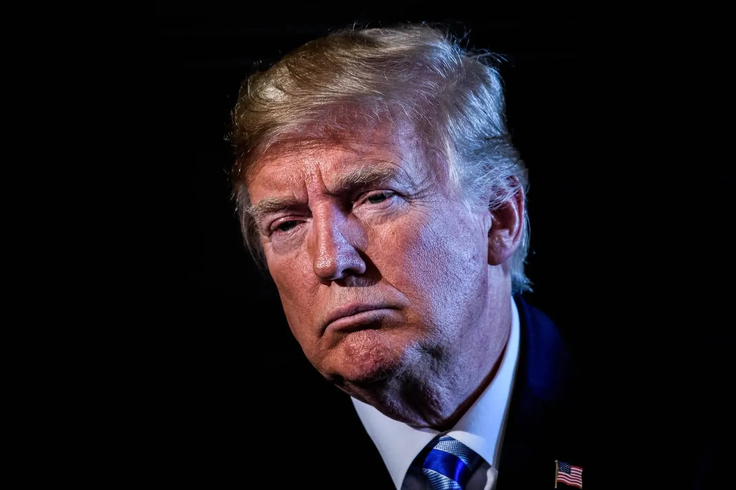 Donald Trump is depicted in this close-up portrait. The image focuses on his face, set against a dark, nondescript background that enhances the details of his expression. His demeanor appears serious and contemplative. The lighting emphasizes the contours of his face, creating shadows that accentuate his features. Donald Trump is wearing a dark suit with a white shirt and a blue striped tie, along with a lapel pin that has the American flag. His hair is styled in his recognizable manner. The image captures a moment of introspection or focus, which is commonly seen in formal portraits of public figures. The overall composition conveys a sense of gravity, often associated with political leadership. There are no visible text elements, logos, or additional contextual objects in the image.