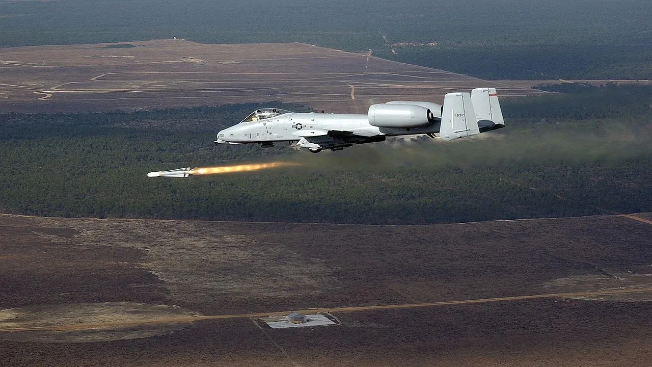 The image depicts an A-10 Thunderbolt II, commonly known as the "Warthog," in flight over a vast landscape of forests and open fields. The aircraft is shown launching a missile, which is visible with a trail of smoke and flames as it moves away from the plane. The A-10 is characterized by its distinctive twin-engine setup and straight-wing design, optimized for close air support missions. It is painted in a light gray color with markings that identify it as part of the U.S. Air Force. The background features a mix of dense forest and open terrain, with a clear sky suggesting daytime conditions. The scene captures the dynamic and powerful nature of the aircraft as it executes a combat maneuver. The lighting is natural, highlighting the details of the plane and the missile's smoke trail. There are no visible people, and the focus remains on the aircraft and its operation.