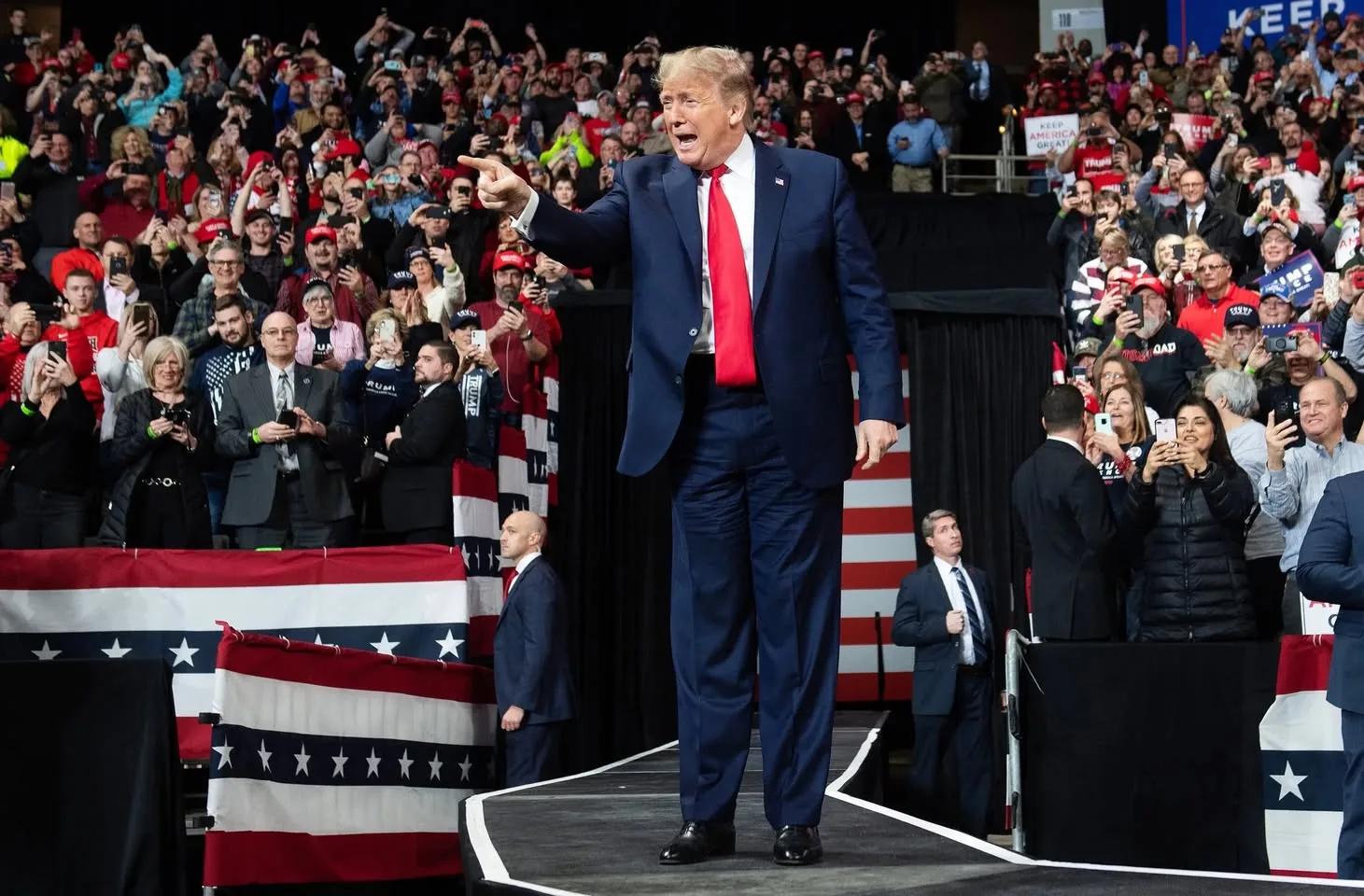 Donald Trump is pictured in this energetic scene, captured at a political rally. He stands confidently on a raised platform, wearing a dark blue suit with a bright red tie, exuding a commanding presence. The background is filled with a dense crowd of supporters, many of whom are capturing the moment on their phones. The atmosphere is lively and vibrant, with attendees wearing red hats and holding signs, some of which bear the phrase "KEEP AMERICA GREAT!"
The setting is an indoor arena, adorned with American flag-themed decorations, adding a patriotic feel to the event. Donald Trump gestures animatedly, pointing into the crowd, suggesting engagement and interaction with the audience. Security personnel are visible nearby, maintaining order as he moves along the stage. The lighting is bright, highlighting Trump and enhancing the visibility of the crowd's enthusiastic expressions. This image vividly conveys the spirited ambiance typical of a political rally.
