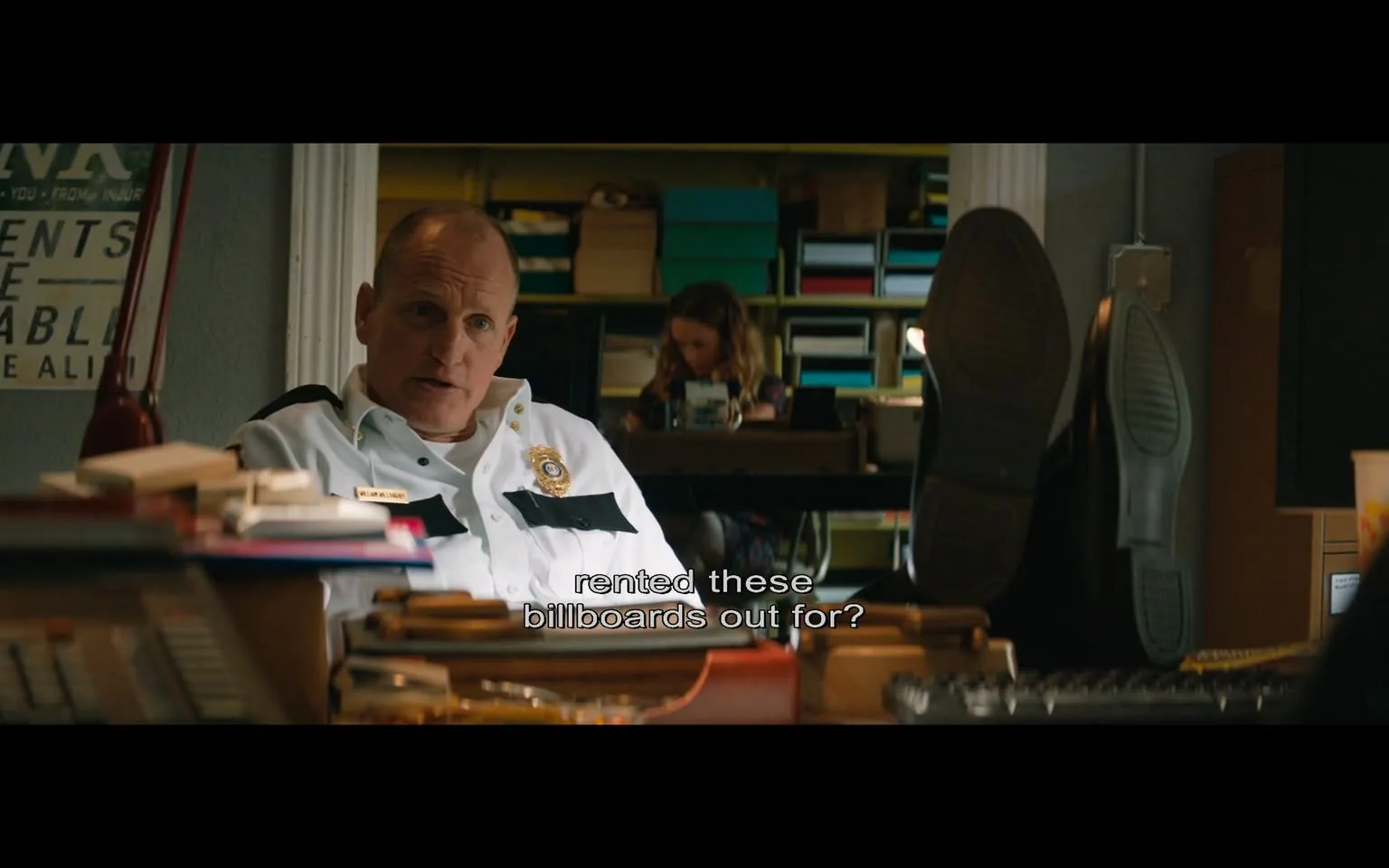 Woody Harrelson is depicted in a scene from the film "3 Billboards Outside Ebbing, Missouri." He is seated in what appears to be an office setting, wearing a white police uniform with black accents and a badge visible on his chest. His posture is relaxed, with his feet up on the desk, suggesting a casual or contemplative moment. The background shows a woman sitting at a desk, focused on a task. The room is filled with shelves holding various colorful boxes and files, indicating a workspace environment. The lighting is warm, adding a natural atmosphere to the scene. A partially visible sign and a red desk lamp contribute to the office’s decor. The subtitle on the screen reads, "rented these billboards out for?", indicating Harrelson's character is engaged in a conversation, possibly questioning or discussing details related to the plot of the film.