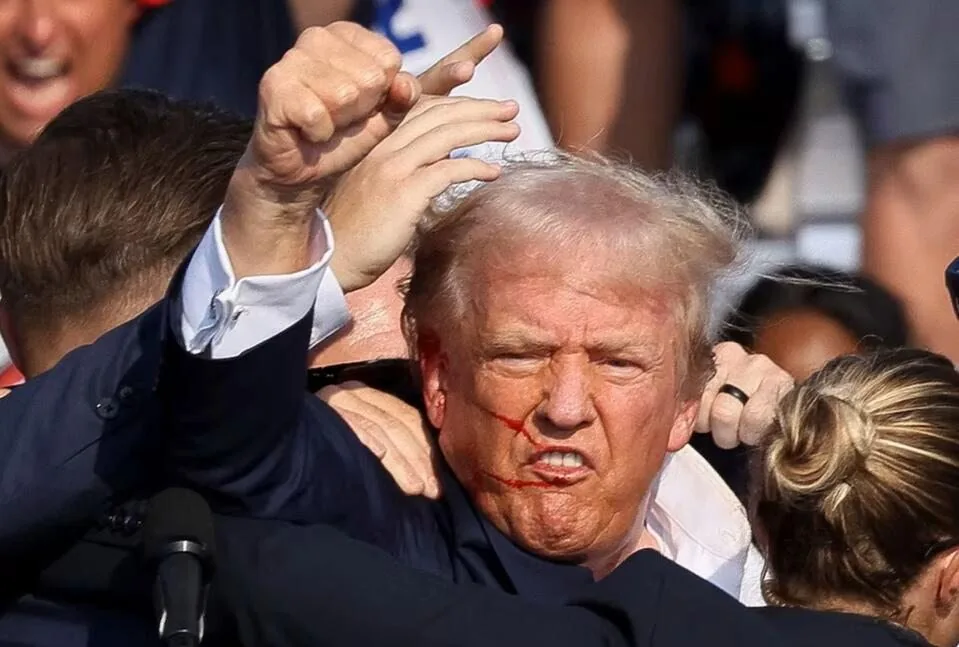 Donald Trump is the central figure in this image. He is surrounded by a crowd, with several hands reaching out towards him. The setting appears to be outdoors during the daytime, as indicated by the lighting and visible crowd in the background. Trump is captured in a moment of intense expression, with his mouth open and brow furrowed, conveying a sense of anger or determination. His hair is disheveled, and there are red marks on his face that give the impression of scratches or smears. He's wearing a dark suit jacket, and his fist is raised in a gesture that suggests defiance or emphasis. The surrounding crowd is animated, with some individuals trying to touch or support him. The overall mood is chaotic and charged, highlighting a moment of tension or protest. The image captures a vivid and dynamic scene, emphasizing the energy and emotion of the moment.