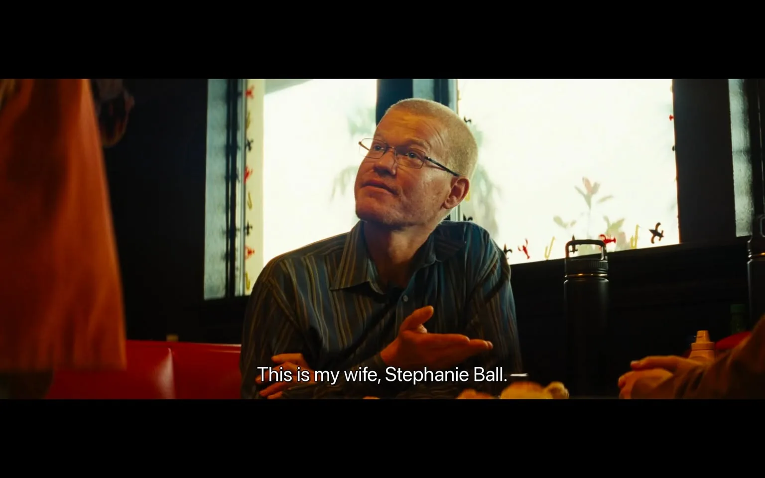 Jesse Plemons is seated in a restaurant setting, captured in a moment of conversation. He is wearing a striped shirt and glasses, and appears engaged, gesturing with one hand while looking towards someone. The background reveals a window with silhouettes of decorative elements, and the lighting suggests it is daytime, casting a soft glow across the scene. The seating is upholstered in red, contributing to a warm and inviting atmosphere. On the table, there are various items, including a dark thermos, indicating a casual dining setting. The subtitle at the bottom of the image reads, "This is my wife, Stephanie Ball," suggesting he is introducing someone. Jesse Plemons' expression is attentive and slightly inquisitive, fitting the context of an introduction or discussion.