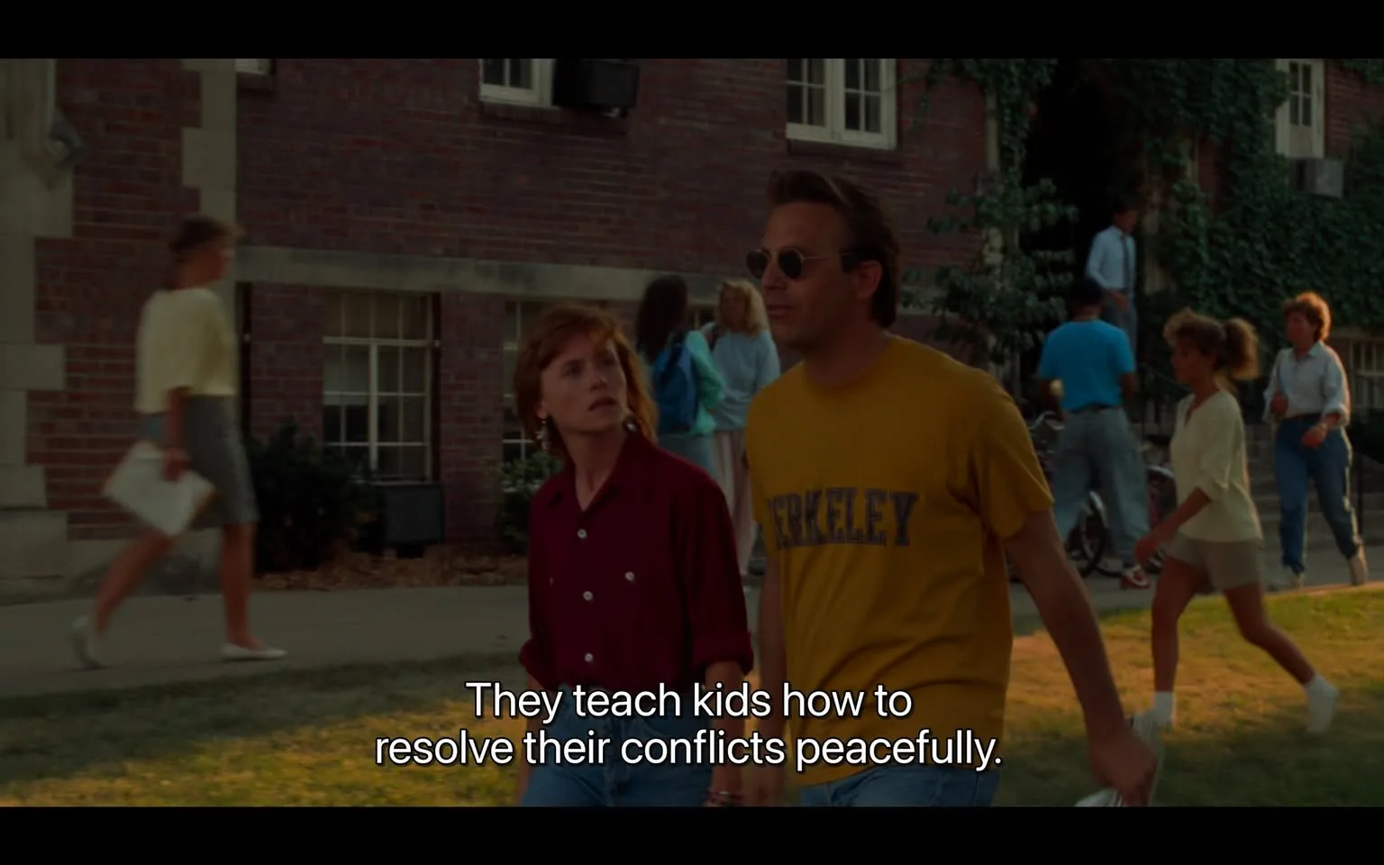 The image depicts a scene from the film "Field of Dreams," featuring Kevin Costner. It shows two people walking on a college campus, surrounded by students. Kevin Costner is wearing a yellow T-shirt with "BERKELEY" printed on it, paired with sunglasses, suggesting a casual and relaxed demeanor. The woman walking beside him, dressed in a red shirt, appears engaged in conversation, looking up at him with curiosity. The subtitle at the bottom of the image reads: "They teach kids how to resolve their conflicts peacefully," indicating a dialogue about education or conflict resolution. The setting is a sunny day, with students casually strolling around, some holding books, which enhances the typical campus atmosphere. The brick building in the background and the presence of greenery add to the collegiate environment. The lighting is bright and natural, reflecting a typical day scene.