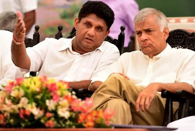 Ranil Wickremasinghe and Sajith Premadasa are seated next to each other at an outdoor event. The scene captures a moment of conversation between the two, with Ranil Wickremasinghe appearing thoughtful and engaged, while Sajith Premadasa gestures as if explaining something. Both are dressed in light-colored shirts, suitable for a formal or semi-formal occasion. They are seated on ornate chairs, indicating a possibly formal or official setting. In the foreground, there is a vibrant bouquet of flowers adding a splash of color to the scene. The background is slightly blurred, suggesting a focus on the two individuals. The lighting is natural, typical of an outdoor event during the day, enhancing the clarity and color of the image. The atmosphere suggests a serious yet cordial discussion, reflective of their roles in a political context.