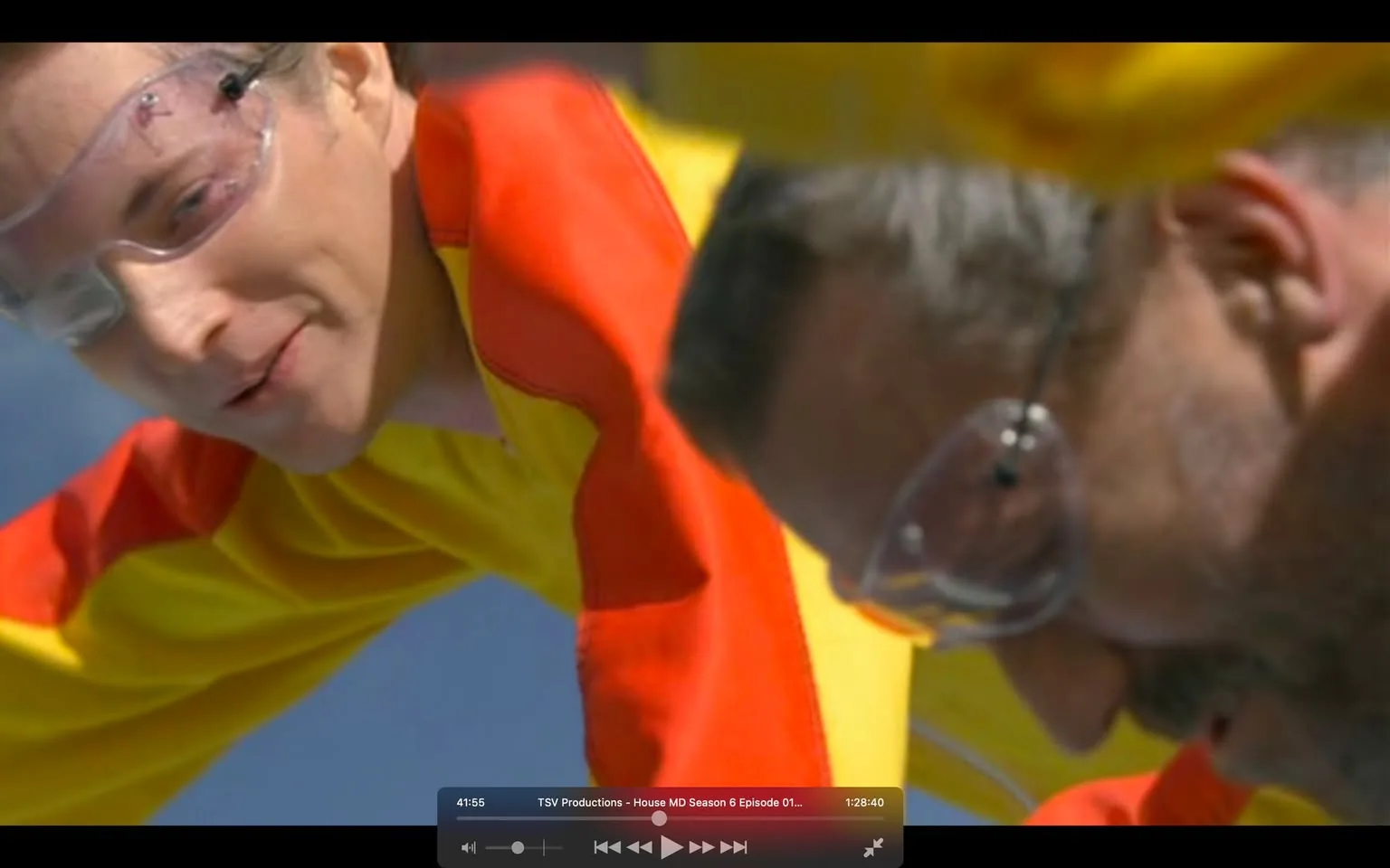 The image is a screenshot from the TV show "House MD," specifically from Season 6, Episode 1. In the scene, two characters are outdoors, engaged in an activity that involves wearing protective goggles and brightly colored suits in red and yellow. One character is smiling slightly, suggesting a sense of enjoyment or camaraderie, while the other appears focused on the task at hand. The vivid colors of their suits contrast with the clear blue sky, highlighting the dynamic and energetic nature of the scene. The close-up angle emphasizes their concentration and involvement in the activity.
The on-screen text at the bottom displays a timestamp, "41:55," and indicates the production company and episode information: "TSV Productions - House MD Season 6 Episode 01," along with the total duration "1:28:40." This enhances the context of the frame as part of a structured episode in a well-known series.