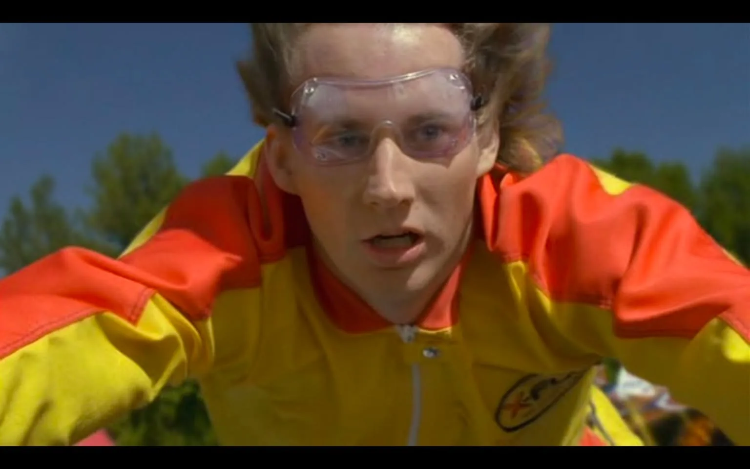 The image is a close-up of a character from "Max Keeble's Big Move," wearing a vibrant yellow and red jumpsuit. The attire is form-fitting and suggests high-speed activity, possibly in a racing or stunt scenario. The character is also wearing clear safety goggles, indicating a need for protection during a fast-paced action scene. The background is a blur of greenery, suggesting an outdoor setting with natural light casting a bright and dynamic atmosphere. The expression on the character's face displays a mix of determination and focus, enhancing the sense of movement and urgency. The composition centers on the character's upper body and face, emphasizing the action-oriented theme of the scene. The style is typical of a live-action film aimed at a younger audience, with bold colors and clear, engaging visuals.