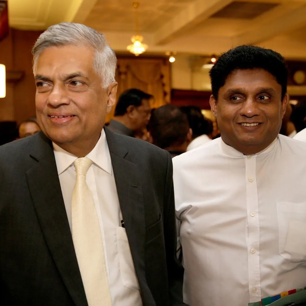 In this image, Ranil Wickremasinghe and Sajith Premadasa are seen together at an indoor event. The setting appears to be formal, with elegant lighting and decor, suggesting a gathering or a ceremony. Ranil Wickremasinghe is on the left, wearing a dark suit with a white shirt and a light-colored tie. He has a subtle smile and is looking to his left. Sajith Premadasa, on the right, is dressed in a traditional white shirt, buttoned to the top, which is common in South Asian formal attire. He is also smiling, looking slightly to his left. The background shows other attendees, indicating a well-attended event, and the lighting is warm, enhancing the formal ambiance. The expressions on their faces suggest a pleasant and cordial interaction.