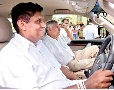 Ranil Wickremasinghe and Sajith Premadasa are seated in a car, with Sajith Premadasa at the steering wheel, appearing to be the driver. Both are dressed in white shirts, contributing to a formal appearance. Sajith Premadasa is smiling, suggesting a relaxed or content mood, while Ranil Wickremasinghe, seated in the passenger seat, appears to be looking out the window, possibly in thought or observation. The interior of the car is visible, showing a dashboard, steering wheel, and part of the vehicle's door. Outside the car, a small group of people is gathered, with one person possibly speaking or gesturing, indicating some interaction or event is taking place. The image is taken during the day, with natural light illuminating the scene. The context suggests it might be related to a political or public event, given the presence of known political figures.