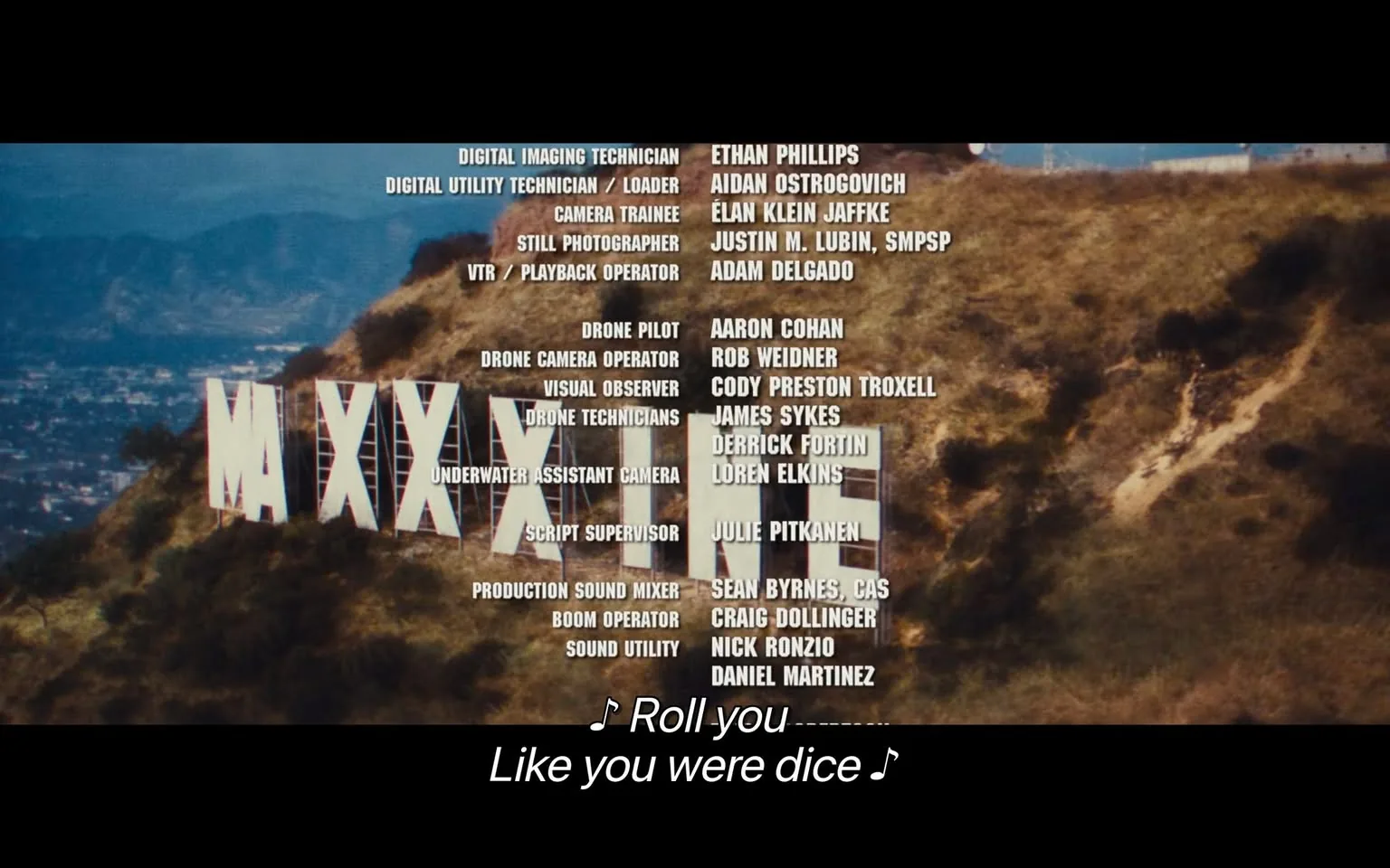 The image is an end credit scene from the film "Maxxxine." It features a large Hollywood-style sign situated on a hillside, spelling out "MAXXXINE" in bold white letters. The background shows a panoramic view of a landscape, likely representing a cityscape, under a clear blue sky.
The credits list various crew members who contributed to the film's production. These include positions such as Digital Imaging Technician, Drone Pilot, and Production Sound Mixer, among others. The names of the individuals who occupied these roles are prominently displayed in white text against the natural backdrop of the hills.
Subtitles at the bottom of the image read, "♪ Roll you / Like you were dice ♪," suggesting a musical or lyrical element is part of the scene. The overall composition conveys a professional production setting with an emphasis on recognizing the technical team behind the film. The scene is brightly lit, emphasizing the grandeur and significance of the sign and the contributions of the crew members.