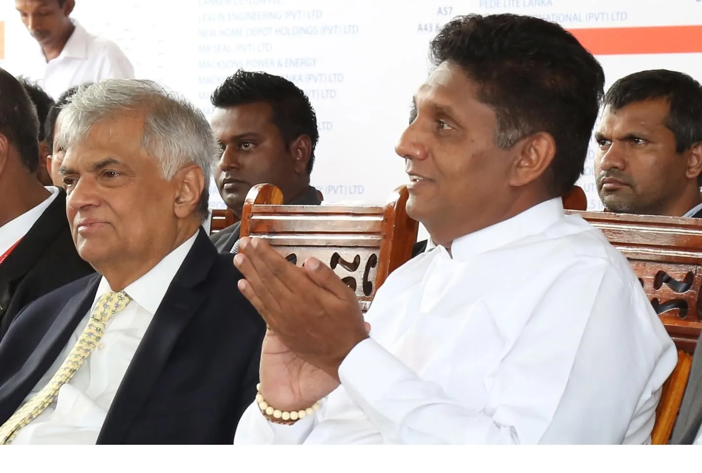 Ranil Wickremasinghe and Sajith Premadasa are depicted in a formal setting, seated next to each other on ornately carved wooden chairs. Ranil Wickremasinghe is dressed in a dark suit with a light-colored tie, looking attentive. Sajith Premadasa, dressed in a traditional white attire, is seen clapping, suggesting engagement in the event. The mood appears formal yet positive. Other individuals in the background are paying attention to the event. The background features a board with text listing various company names, indicative of a corporate or official function. The lighting is bright, typical of an indoor daytime event. The focus on the two individuals suggests their prominence in the gathering.