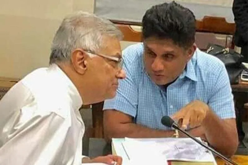 ```
Ranil Wickremasinghe and Sajith Premadasa are engaged in a focused conversation in this indoor setting. Ranil Wickremasinghe, with gray hair and wearing glasses, is dressed in a formal white shirt. He is sitting on the left, facing Sajith Premadasa, who is wearing a light blue checkered shirt. Sajith is leaning slightly forward, pointing towards a document on the table, indicating a serious discussion. The setting appears to be a meeting room, with papers and a microphone in front of them, suggesting a formal or political context. The lighting is bright and even, emphasizing the professional atmosphere. The background shows a typical office environment with a desk and chairs, adding to the formal tone of the scene.