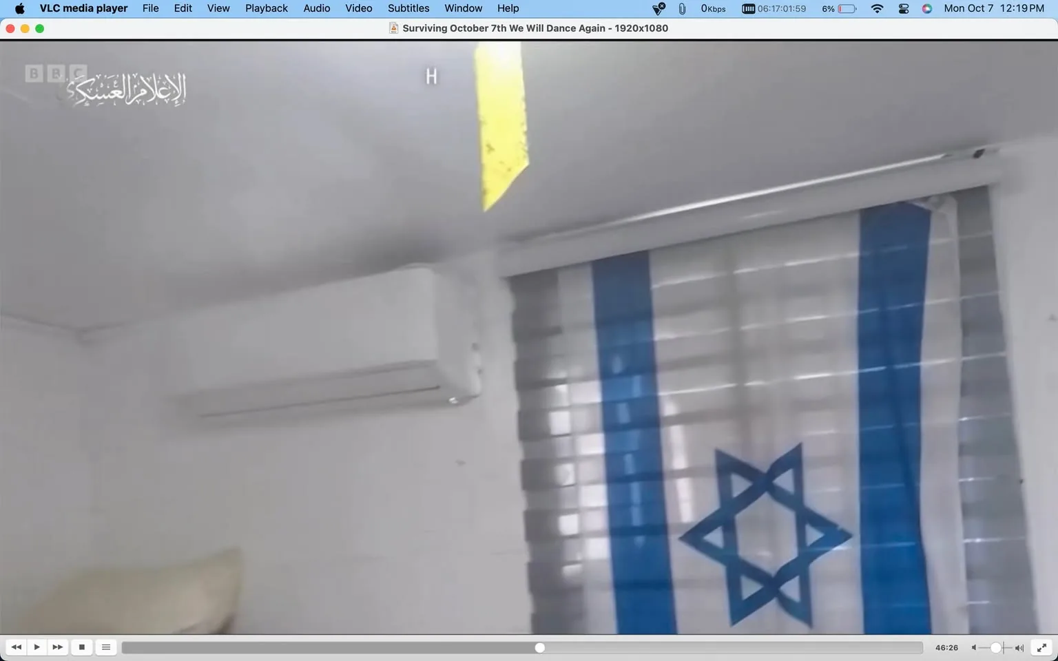 The screenshot shows a paused frame from a video being played on VLC media player, titled "Surviving October 7th We Will Dance Again - 1920x1080." The interface of the media player is visible at the top and bottom of the screen. The setting appears to be a room with white walls. An Israeli flag is prominently displayed in the background, partially covering a window with horizontal blinds. The flag, with its blue Star of David, is a key focal point in the image. To the left of the flag, an air conditioning unit is mounted on the wall. The lighting is bright, suggesting daytime, and there’s a yellow strip of paper or tape hanging from the ceiling. The text "BBC" is seen in the top left corner, along with Arabic script reading "الإعلام العسكري" (Al-I'lam Al-'Askari), likely indicating military media. The atmosphere is neutral, focused on documenting or reporting specific events related to the Israeli context.