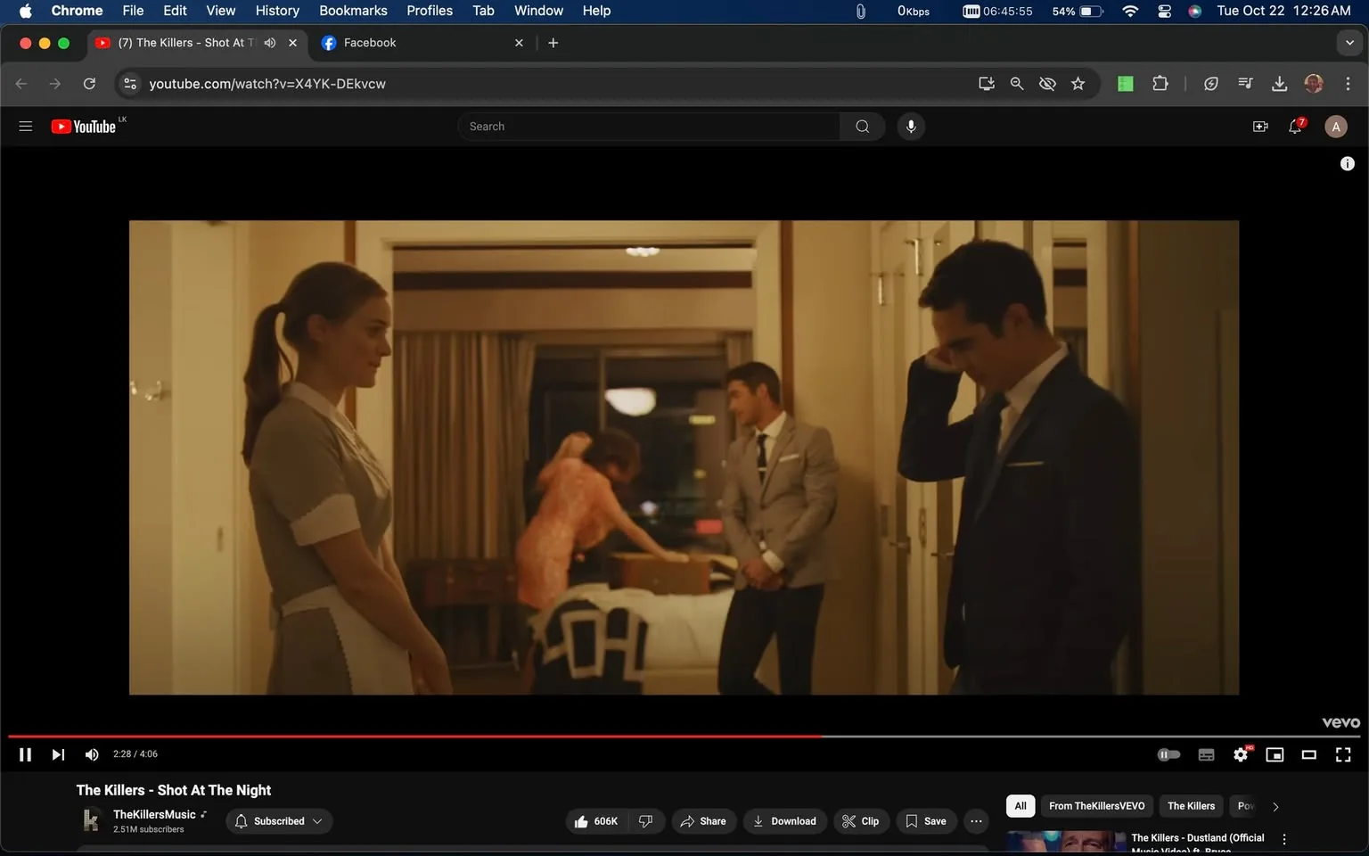 The scene is from the music video "The Killers - Shot At The Night," showcasing an indoor setting within a hotel room. In the foreground, a woman in a maid’s uniform stands composed, with her hair neatly tied back, indicating she might be part of the hotel staff. Next to her, a man in a suit stands with a contemplative pose, touching his temple, suggesting he might be deep in thought or stressed. In the background, another man, also in a suit, leans casually against the wall, watching a woman in a bright dress who seems to be bustling around the room. The lighting is warm, casting a cozy, intimate glow over the scene, enhancing the mood of introspection and quiet tension.
- Title: "The Killers - Shot At The Night"
- Channel / profile: "TheKillersMusic"
- Site / app: "YouTube"