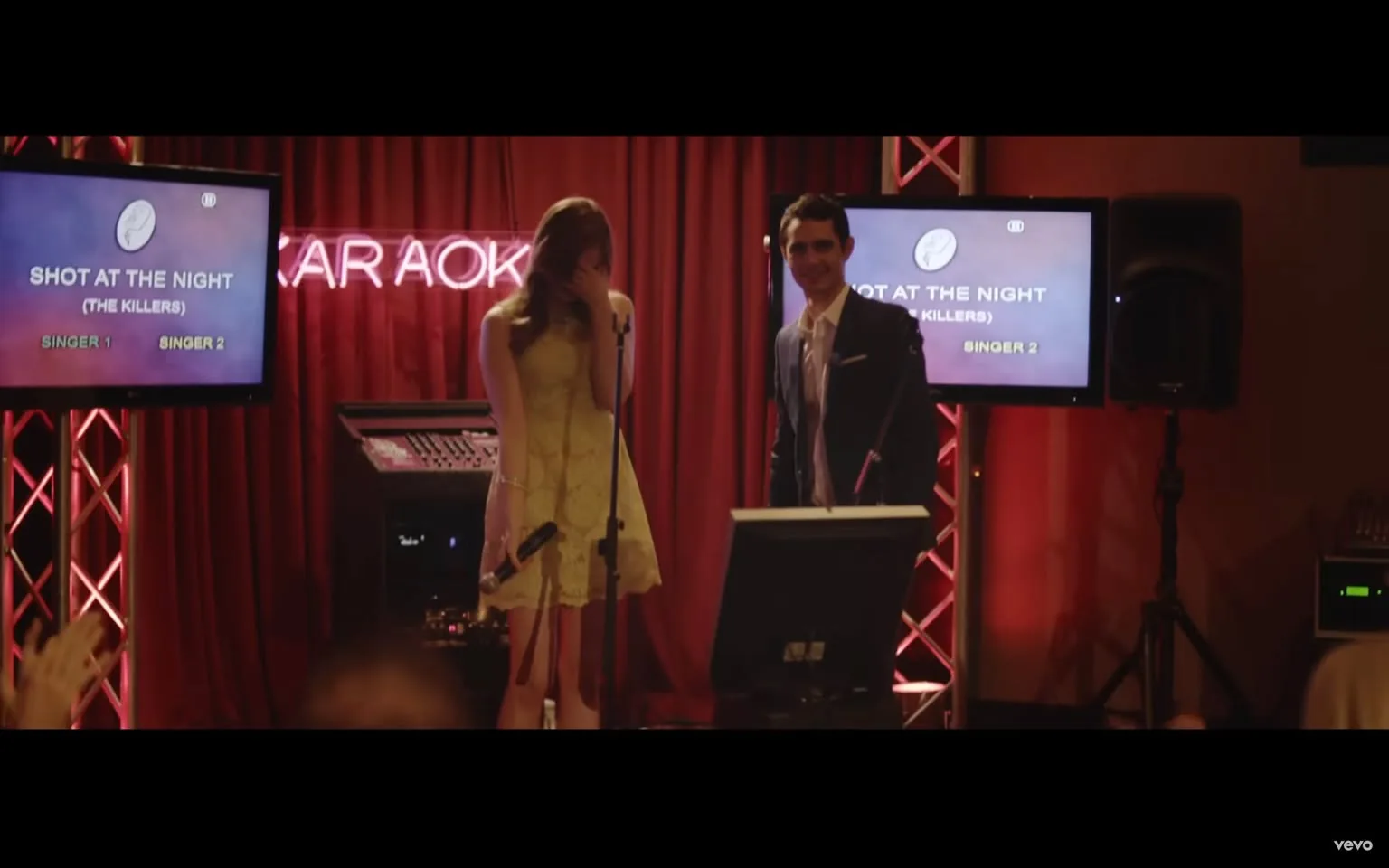 This image is a frame from the music video "Shot at the Night" by The Killers, as indicated by the hint. The scene takes place in a karaoke bar setting, with a red curtain backdrop and neon lighting that spells out "KARAOKE" in the background. A man and woman are standing at the front, both holding microphones, suggesting they are about to perform a duet. The woman, dressed in a light yellow dress, is covering her mouth with one hand, perhaps out of nervousness or laughter. The man is wearing a dark suit and is smiling confidently. The lighting is warm, casting a cozy glow over the scene. Two screens flanking the performers display the song title "SHOT AT THE NIGHT" and the artist "(THE KILLERS)," along with placeholders for "SINGER 1" and "SINGER 2." The audience's hands are visible in the foreground, indicating engagement and anticipation. The overall atmosphere is lively and intimate, characteristic of a karaoke event.
