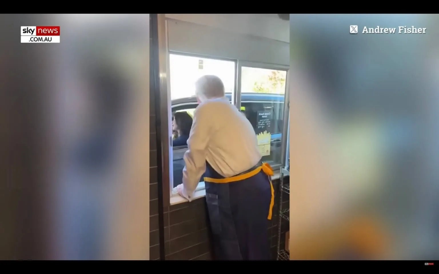 1) Rich description (8–12 sentences): In the image, a man is leaning out of a drive-thru window, engaging with a customer in a vehicle. Based on the hint, this scene includes Donald Trump. The setting is a fast-food restaurant's drive-thru, with an indoor view from behind the counter. The man, wearing a white shirt and a blue apron with a yellow tie, is leaning forward, suggesting an active interaction. The vehicle's driver appears attentive, looking up towards the window. The atmosphere seems casual and dynamic, typical of a drive-thru exchange. The lighting is bright, indicating daytime or a well-lit indoor environment. The video frame includes branding from "Sky News Australia," with the logo visible in the upper left corner. The name "Andrew Fisher" is displayed in the top right, suggesting content attribution.
2) On-screen text block (only include lines that have real text; if a field is unknown or blank, omit that line entirely):
- Site / app: "Sky News Australia"
- Channel / profile: "Andrew Fisher"
- Key OCR Lines:
- "sky news"
- "COM.AU"