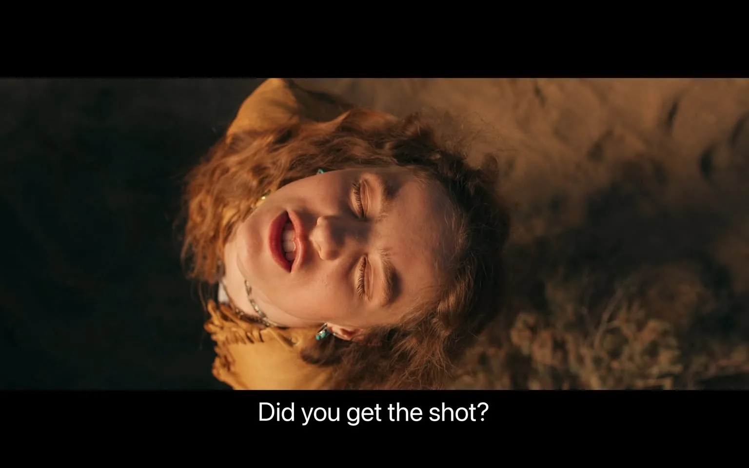 In this video frame, a young woman is captured in a close-up shot with her head tilted back, suggesting she may be lying on the ground. Her eyes are closed as she speaks, and her expression appears intense or in the midst of exertion. Her curly hair is visible, and she is wearing a yellow top. The background is blurred, possibly depicting a sandy or earthy terrain, suggesting an outdoor setting. The lighting is warm, which highlights her facial features and hair. The subtitle at the bottom reads, "Did you get the shot?" indicating she might be involved in some kind of dramatic or pivotal moment. The scene's overall tone hints at urgency or anticipation.
