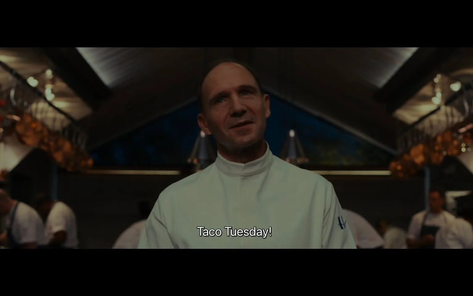 Ralph Fiennes is featured in this scene from the film "The Menu." He appears to be in a professional kitchen setting, wearing a white chef's uniform, which emphasizes his role as a chef in the movie. The atmosphere has a warm, dimly lit ambiance, suggesting an evening or night time. In the background, other chefs are visible, possibly preparing food, adding to the busy and focused environment typical of a high-end kitchen. The architecture of the kitchen includes high ceilings and elegant lighting fixtures. Ralph Fiennes has a slight smile on his face, and his posture is relaxed yet authoritative, suggesting confidence and control in his environment. The composition of the shot centers on him, highlighting his importance in the scene. The subtitle “Taco Tuesday!” is displayed, hinting at a thematic or humorous element within this part of the film.
- Captions / subtitles: "Taco Tuesday!"