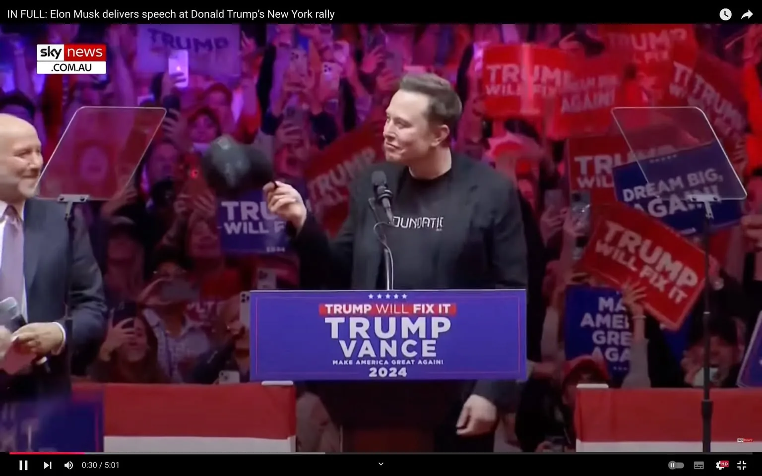 1) Rich description: The image captures a lively scene at a political rally, with Elon Musk standing at a podium. The podium is adorned with a sign reading "TRUMP WILL FIX IT," followed by "TRUMP VANCE" and "MAKE AMERICA GREAT AGAIN! 2024." Elon Musk is wearing a black jacket over a shirt with visible text, gesturing and holding a hat in one hand. The background is filled with a vibrant, cheering crowd holding signs that say "TRUMP WILL FIX IT" and "MAKE AMERICA GREAT AGAIN!" The atmosphere is energetic, with bright purple and red lighting casting a colorful hue over the crowd. The setting suggests a significant political event, with clear branding and messaging aimed at promoting a political campaign. A man in a suit stands nearby, appearing to be engaged in the proceedings, while the audience captures the moment on their smartphones.
2) On-screen text block:
- Title: "IN FULL: Elon Musk delivers speech at Donald Trump's New York rally"
- Channel / profile: "sky news"
- Site / app: "YouTube"