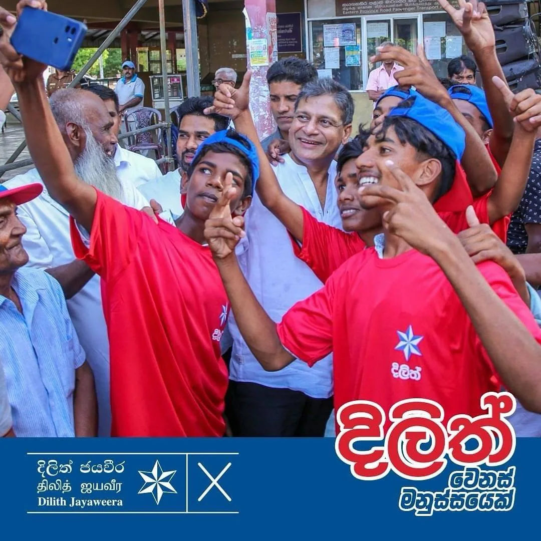 ```
1) Rich description: The image shows a lively group of people gathered outdoors, taking a selfie. At the center, a man, identified as Dilith Jayaweera, is smiling warmly amidst a crowd of young people wearing bright red shirts and blue caps. The shirts feature a white star symbol, and the overall mood is cheerful and energetic. One of the young individuals extends their arm to capture the moment on a smartphone. The setting appears to be a public area, possibly a street or square, with a backdrop of a building featuring posters and notices. The lighting is natural and suggests a sunny day, enhancing the vibrant colors of the attire. The group seems to be in a celebratory or promotional mood, possibly indicative of a campaign or public event. The bottom of the image includes a blue banner with text in both Sinhalese and English, prominently displaying "Dilith Jayaweera" and a star symbol.
2) On-screen text block:
- Key OCR Lines: "දියත්", "Dilith Jayaweera"