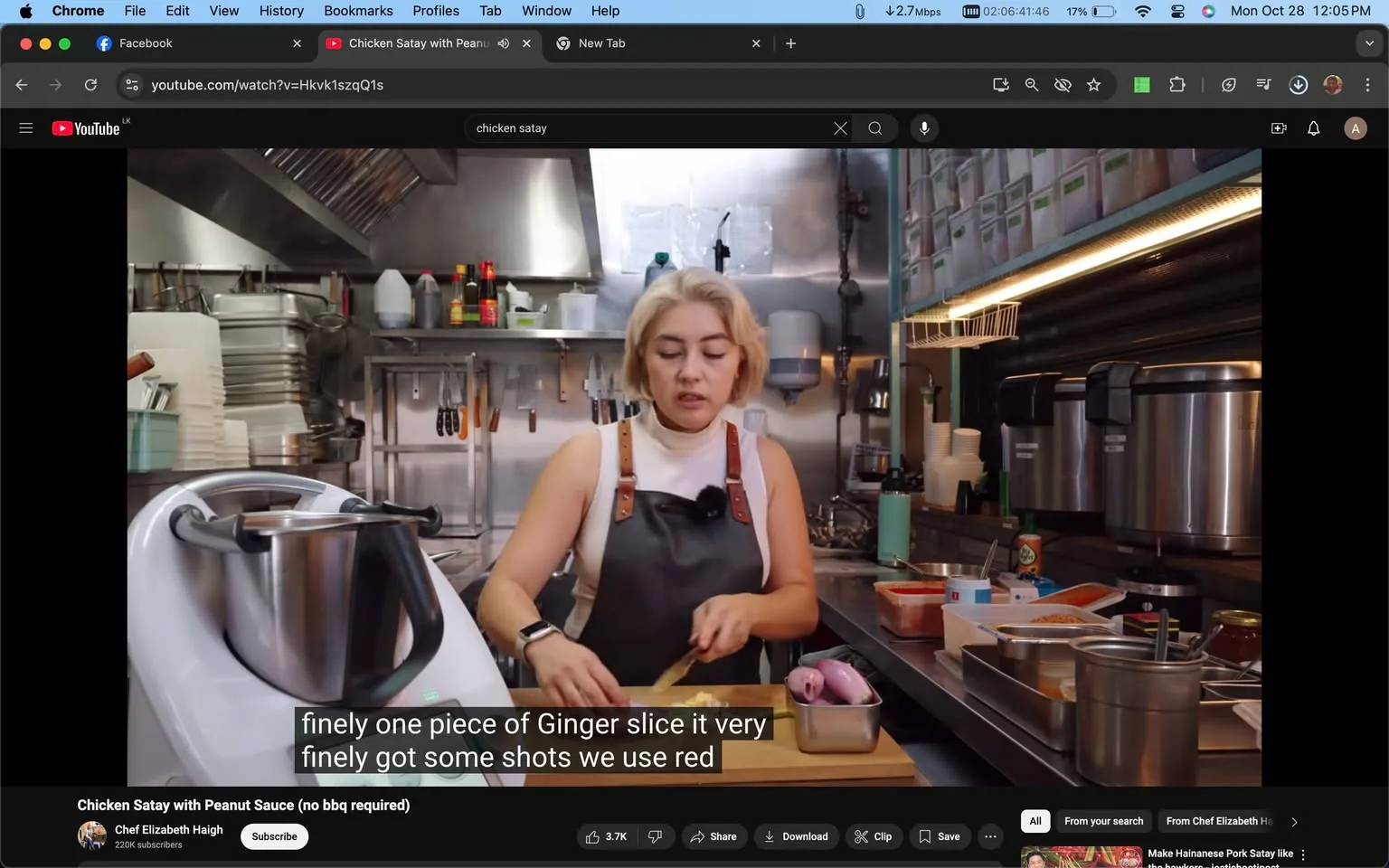 ---
The image is a screenshot from a YouTube video titled "Chicken Satay with Peanut Sauce (no bbq required)" by Chef Elizabeth Haigh. The video is paused at a moment where Chef Elizabeth is in a professional kitchen, wearing a black apron over a white sleeveless top. She is chopping ingredients on a wooden cutting board, with a focus on finely slicing ginger. Her expression is attentive and focused on the task at hand. The kitchen is well-equipped, featuring various utensils, stacked plates, and industrial kitchen appliances. A large blender or food processor is visible on the left side of the counter, suggesting the preparation of a sauce or marinade. The background is filled with organized kitchen supplies, enhancing the professional cooking environment. The lighting in the kitchen is bright, highlighting the preparation area. The screenshot also displays subtitles at the bottom, providing instructions on slicing ginger. The YouTube interface is visible, with the video title and details about the channel shown below the video frame.