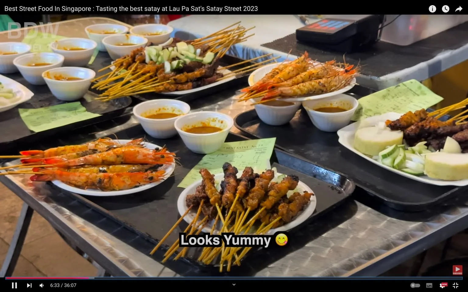 The image is a scene from a video titled "Best Street Food In Singapore: Tasting the best satay at Lau Pa Sat's Satay Street 2023" on the BDW Travel TV channel on YouTube. This video frame showcases a vibrant display of street food, specifically satay, at Lau Pa Sat's Satay Street in Singapore. In the foreground, several plates are filled with skewers of grilled meat and prawns, surrounded by small bowls of dipping sauce. The satay is arranged neatly on the plates, garnished with sliced cucumbers and onions, and accompanied by rice cake pieces known as "ketupat."
The skewers are golden and charred, indicating they are freshly grilled, while the prawns have a bright orange hue, adding to the appetizing appeal. The food is set on a metal table or serving counter, typical of a bustling food market environment. Small green papers, possibly order tickets, are visible among the dishes. The lighting is bright, highlighting the vivid colors of the food and creating an inviting atmosphere. The subtitle "Looks Yummy 😊" is visible at the bottom, reflecting the enticing nature of the display.