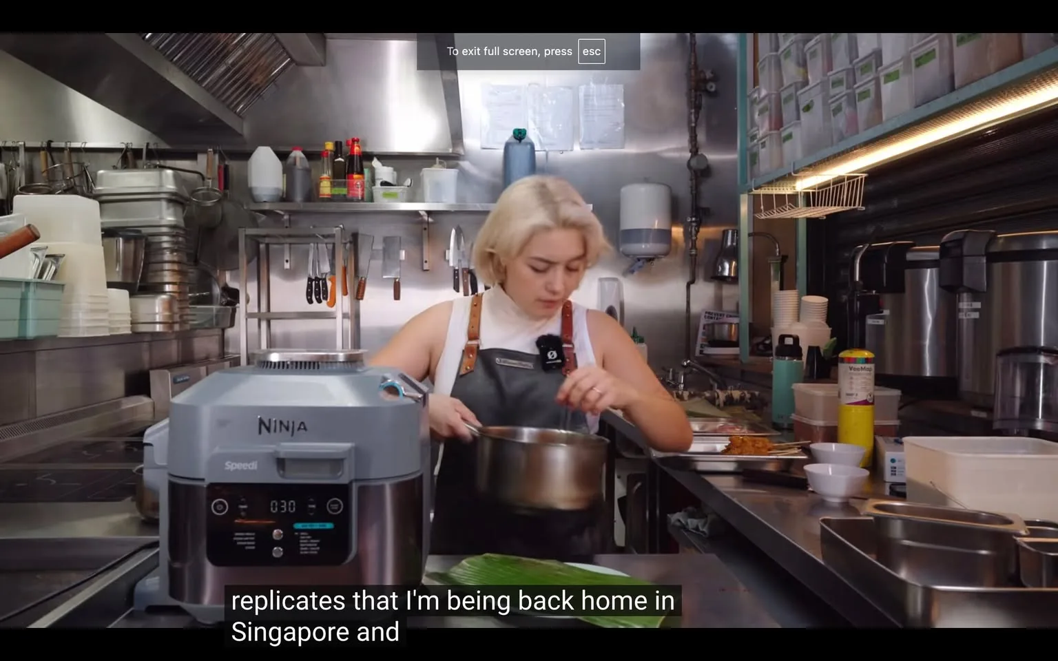 The image depicts a kitchen scene, likely from a cooking video or show. A person with light-colored hair is in focus, wearing a white top and a dark apron. They are engaged in preparing food, holding a pot while standing at a countertop. The kitchen is equipped with various tools and appliances; a Ninja Speedi appliance is prominently displayed in the foreground. In the background, there are shelves with jars, utensils hanging on hooks, and bottles of sauces. The overall atmosphere is busy and professional, indicative of a commercial kitchen. The lighting is bright, highlighting the stainless steel surfaces and various kitchen items.
- Captions / subtitles: "replicates that I'm being back home in Singapore and"