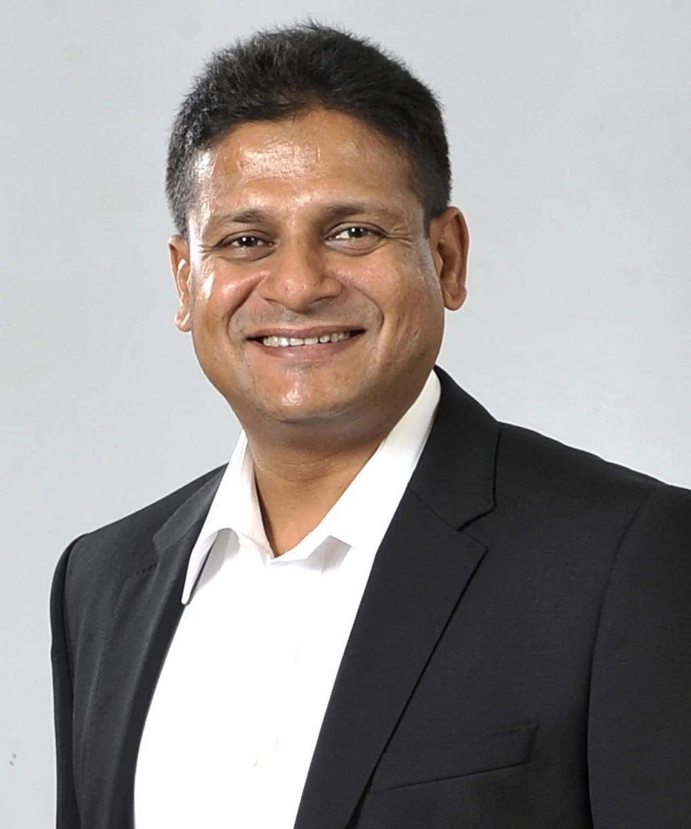 ```
Dilith Jayaweera is depicted in a professional portrait photograph. He is wearing a dark suit jacket over a crisp white shirt, exuding a formal and polished appearance. The attire suggests a business or professional context. His expression is friendly and confident, featuring a broad smile that conveys approachability. The background is plain and light-colored, likely a studio setting, which keeps the focus on him. The lighting is even and soft, highlighting his features and giving a clean, professional look to the image. This photograph could be used for corporate profiles, media publications, or professional networking platforms. There are no visible objects or text in the image, ensuring that Dilith Jayaweera is the main focus.