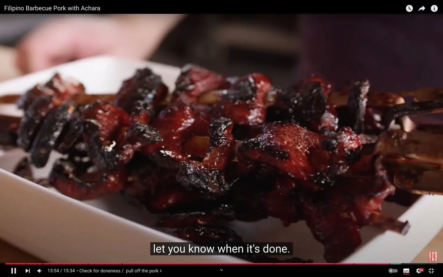 The video frame features a close-up of a plate of Filipino barbecue pork, which is known for its rich red glaze and slightly charred edges, giving it a caramelized appearance. The pork is skewered and served on a white plate, highlighting the vibrant colors of the dish. The setting appears to be a kitchen, suggested by the presence of blurred utensils and a hand in the background, possibly indicating someone preparing or presenting the food. The lighting is warm, enhancing the appetizing look of the barbecue.
The title of the video, "Filipino Barbecue Pork with Achara," is prominently displayed at the top, suggesting the dish being focused on. Subtitles at the bottom read, "let you know when it's done," indicating a step in the cooking process. The scene conveys a homely and inviting atmosphere, typical of cooking shows that focus on sharing recipes and culinary techniques.
- Title: "Filipino Barbecue Pork with Achara"
- Site / app: "YouTube"
- Captions / subtitles: "let you know when it's done."