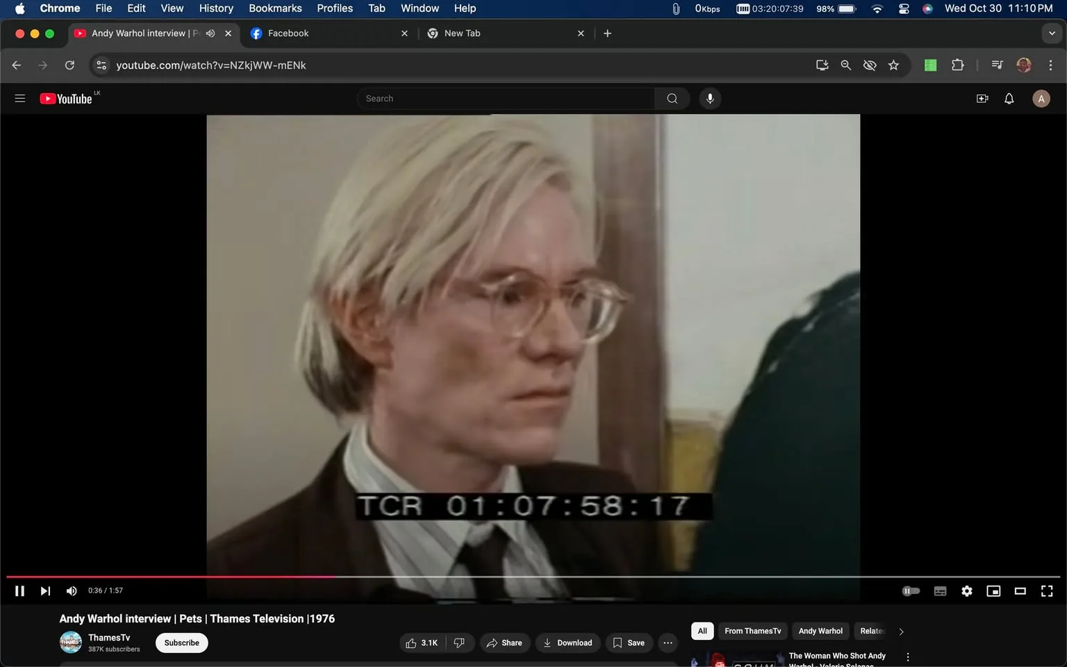 Andy Warhol is featured in this video frame from a 1976 interview titled "Andy Warhol interview | Pets | Thames Television." The setting is indoors, possibly a studio or an interview room. Warhol is dressed in a formal dark brown suit, a white collared shirt, and a striped tie, reflecting the fashion of the era. He wears large, transparent glasses, and his iconic platinum blonde hair is styled in a neat, straight fashion. His expression appears serious and contemplative. The lighting is soft, casting gentle shadows on his face, which adds to the subdued atmosphere of the scene. The timestamp "TCR 01:07:58:17" is visible at the bottom of the frame, indicating the timecode in the video. The video is hosted on YouTube, under the channel "ThamesTV." The visual composition focuses on Warhol's upper body, capturing his thoughtful demeanor during the interview.