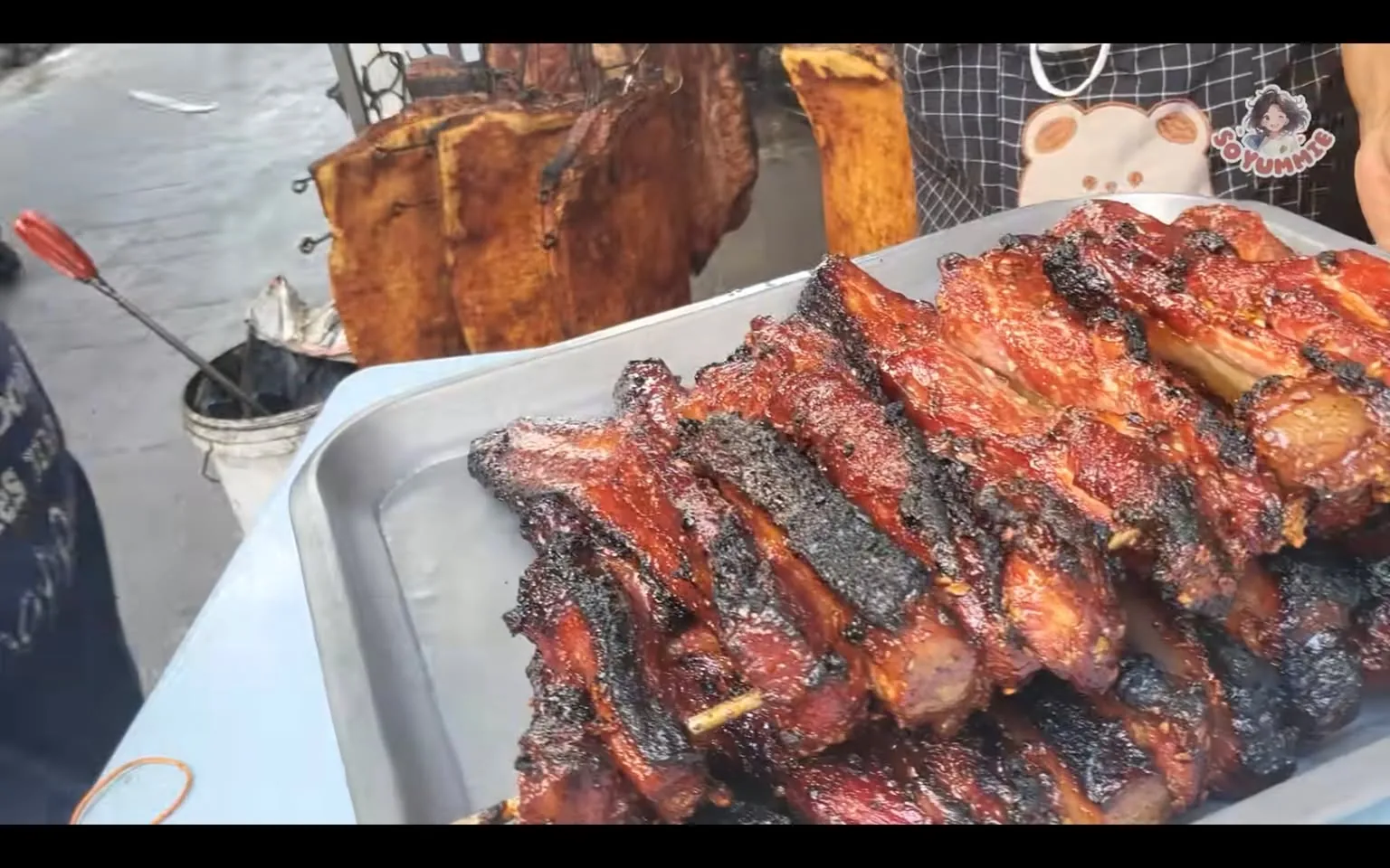 The image depicts a busy street food stall setting, likely outdoors. In the foreground, there is a metal tray filled with skewers of grilled meat, which appears to be perfectly charred and glazed, giving it a shiny and appetizing appearance. The meat is stacked neatly, showcasing its juicy texture and smoky flavor. In the background, larger pieces of meat are hanging, possibly for additional grilling or as a visual display to attract customers.
The vendor, partially visible, is wearing an apron with a bear design, adding a whimsical touch to the scene. The apron suggests a playful and inviting atmosphere typical of street food markets. The environment suggests a bustling area with other stalls or activities nearby, indicated by the concrete pavement and additional cooking equipment visible around the setup. The lighting is natural, suggesting daytime, which enhances the vibrant colors of the grilled meat, making it visually appealing.
- Channel / profile: "Soyumike"