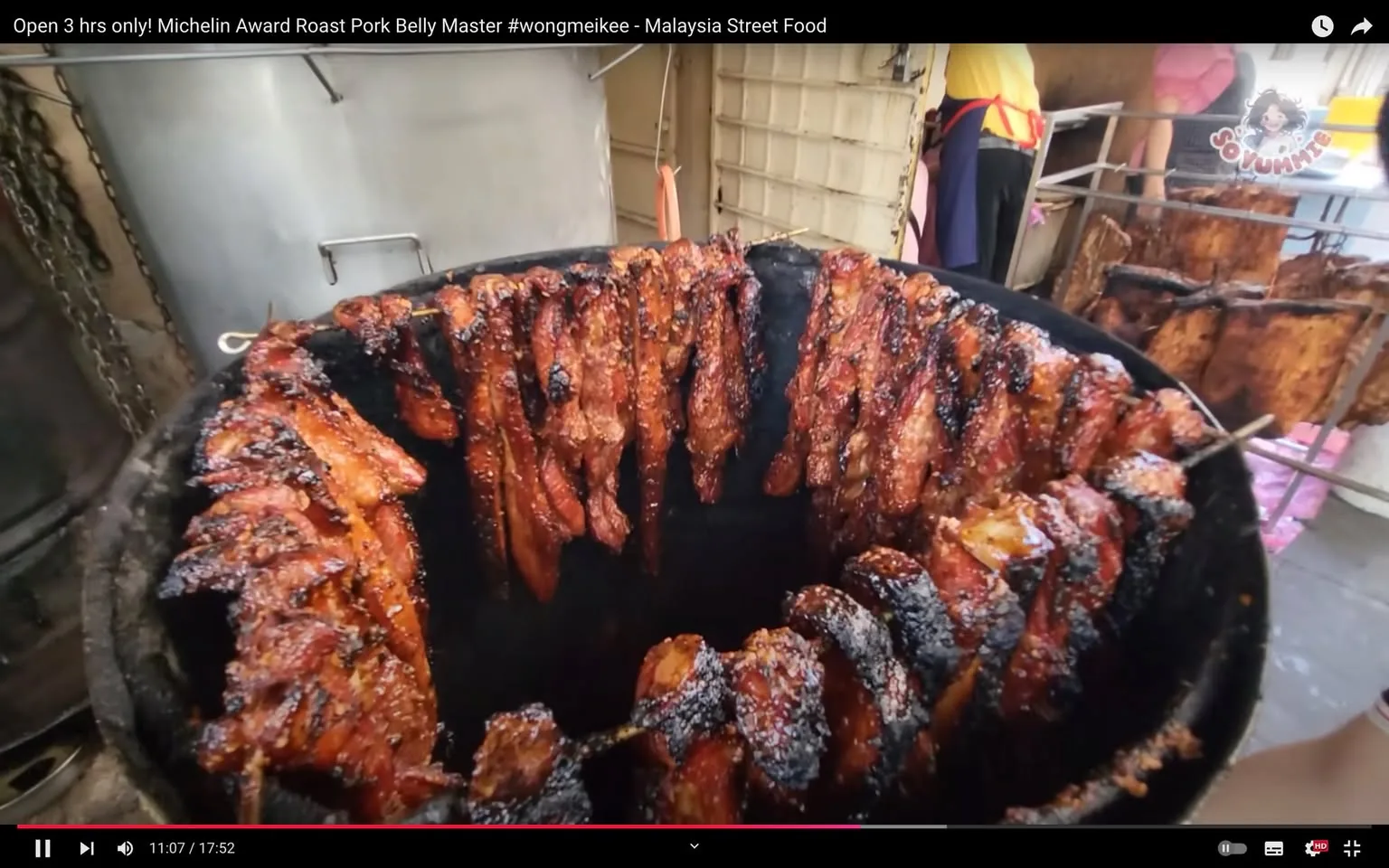 The image is a video frame showcasing a bustling street food scene, highlighting the preparation of roast pork belly. The setting is outdoors, likely at a market or a street food stall in Malaysia. In the center of the frame, numerous skewers of pork belly are hung vertically around the interior of a large roasting drum, each piece glistening with a caramelized, golden-brown glaze, indicating they are being roasted to perfection. The texture of the pork suggests it is both juicy and crispy, with charred edges that promise a smoky flavor.
In the background, there are glimpses of the bustling activity behind the cooking area, where a person is partially visible, wearing a yellow shirt and a red apron, engaged in food preparation tasks. The environment is lively and informal, characteristic of a street food setting, with the presence of metal hooks and chains supporting the cooking apparatus. The lighting is natural, enhancing the rich colors of the roasted pork against the darker tones of the roasting drum.
The video is titled "Open 3 hrs only! Michelin Award Roast Pork Belly Master #wongmeikee - Malaysia Street Food," suggesting the featured food item has received acclaim, perhaps from a Michelin-awarded vendor. The frame is from a video associated with the channel "So Yummy," which likely focuses on diverse and delectable culinary experiences. The scene captures the essence of street food culture, where the art of roasting is both a visual and sensory delight.