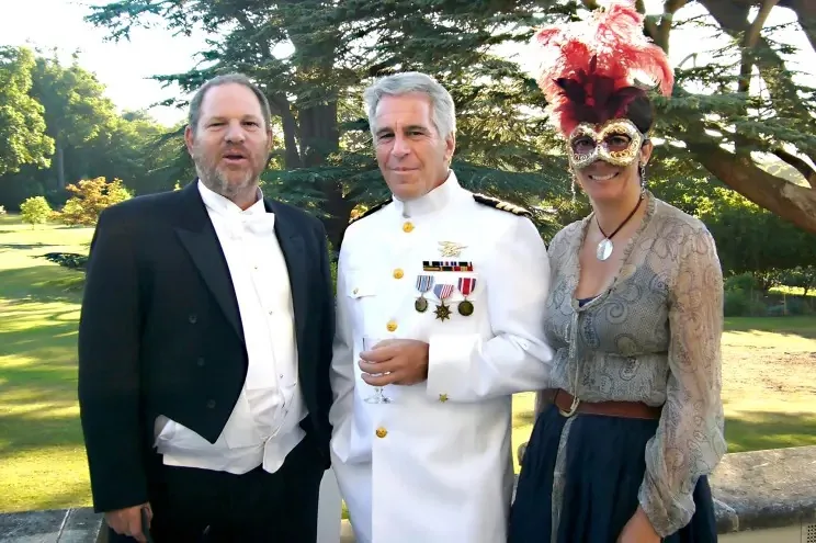 Jeffrey Epstein, Harvey Weinstein, and Ghislaine Maxwell are pictured together outdoors during a formal event. Epstein stands in the center wearing a white naval uniform adorned with medals, suggesting a costume or themed gathering. He holds a drink and has a composed expression. On his right, Harvey Weinstein is dressed in a traditional black tuxedo with a white bow tie, his posture relaxed and demeanor casual. To Epstein's left, Ghislaine Maxwell wears a detailed masquerade-style mask with red feathers and a patterned top, contributing to the festive and costumed atmosphere. The background features lush greenery and towering trees, indicating a garden or estate setting in daylight. The overall mood is formal yet celebratory, with the individuals appearing to be part of a special occasion or themed event. Lighting is natural, with the sun casting soft shadows and highlighting the vibrant colors of the attire and setting.