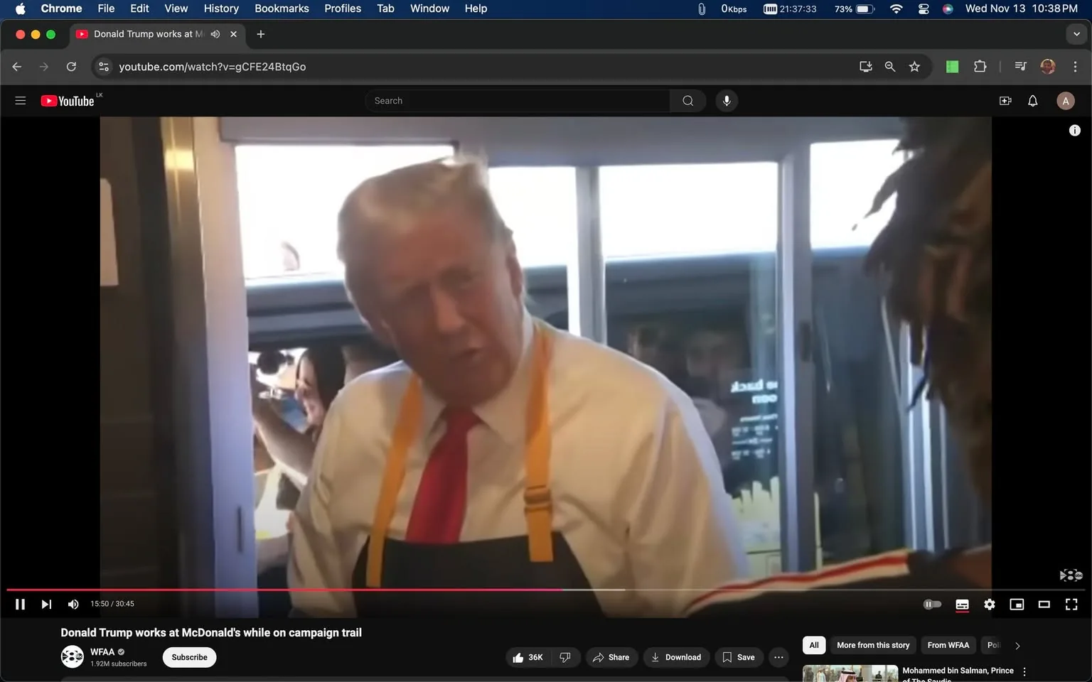 The image is a screenshot from a YouTube video titled "Donald Trump works at McDonald's while on campaign trail." It features Donald Trump inside a McDonald's, wearing a white shirt, a red tie, and a black apron with yellow straps, appearing to engage with someone off-camera. The setting is inside a fast-food restaurant, with the drive-thru window visible in the background. The lighting is bright, capturing the indoor ambiance of a typical McDonald's. Behind Donald Trump, a person is visible through the window, possibly taking a photo or video with a smartphone. The atmosphere seems lively and informal, indicative of a campaign trail event where Donald Trump is interacting directly with people. The YouTube channel hosting the video is "WFAA."
- Title: "Donald Trump works at McDonald's while on campaign trail"
- Channel / profile: "WFAA"
- Site / app: "YouTube"