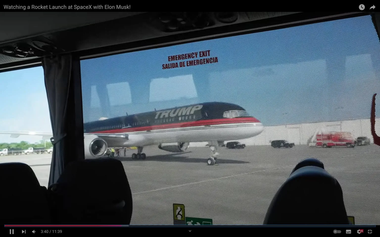 The image is a screenshot from a video titled "Watching a Rocket Launch at SpaceX with Elon Musk!" It shows a view from inside a vehicle, looking out at an airport runway where a plane with the name "TRUMP" painted on its side is stationed. The plane features a sleek design with a black, white, and red color scheme. The setting is daytime, with clear skies indicating good weather. 

The view through the window is partially obscured by a mesh screen, with the text "EMERGENCY EXIT" and "SALIDA DE EMERGENCIA" visible on the glass. The interior of the vehicle includes seat backs, suggesting it might be a bus or similar transport. There's a sense of movement, possibly indicating the vehicle is in transit across the airport tarmac. The overall mood of the image is one of anticipation and transit, fitting the theme of a journey or event at SpaceX.