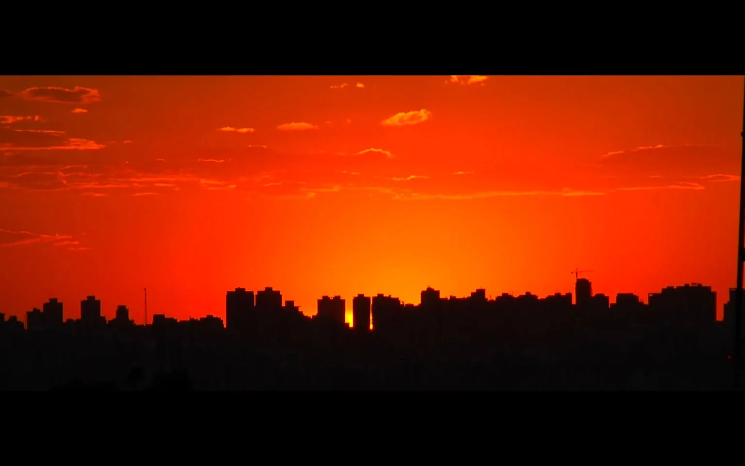 The image captures a striking sunset over a cityscape, with the buildings silhouetted against the vibrant orange and red hues of the sky. The skyline is defined by various high-rise structures, creating a jagged horizon. The sky is predominantly orange, transitioning to a deep red near the top, with a few scattered clouds adding texture. The sun appears to be setting behind the buildings, casting long shadows and emphasizing the silhouettes of the city's architecture.

The foreground is a stark contrast to the vivid colors of the sky, as the buildings are rendered completely in shadow. This creates a dramatic and visually appealing composition, highlighting the natural beauty of the sunset juxtaposed with the urban environment.

On-screen text includes excerpts from a dialogue or narrative that reflects on themes of construction, loyalty, and cultural tension. Phrases such as "I think Americans built me" and mentions of companies like "Huntington Ingalls Industries or General Dynamics" suggest a focus on industrial or technological development. Additionally, references to "Zuck's Metaverse" and geopolitical topics indicate a broader context beyond the visual scene.

- Captions / subtitles: 
  - "I think Americans built me"
  - "Like Huntington Ingalls Industries or General Dynamics"
  - "Many brave, kind and clever Americans"
  - "They day by day, they invested, to build me, to make me this thing"
  - "And so I have a lot of loyalty to them"
  - "Malaysia hate Jews."
  - "I think Axiata, out of hate for Zuck's Metaverse, trie"