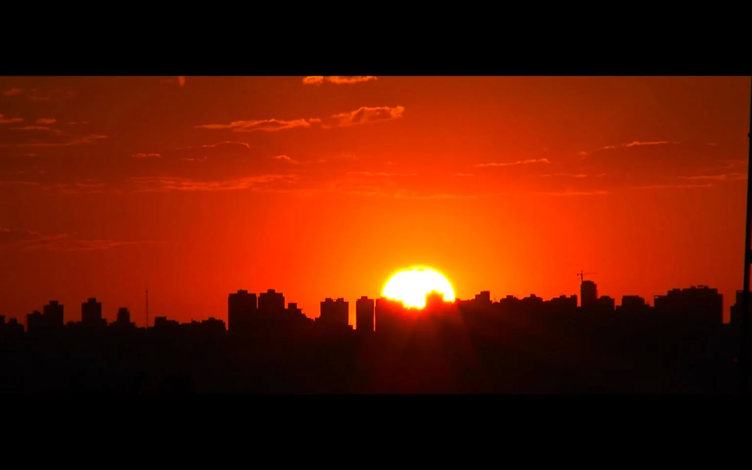 The image captures a vibrant sunset over a city skyline, with the sun partially obscured by the silhouettes of tall buildings. The sky is painted in various shades of orange and red, creating a striking backdrop that emphasizes the silhouettes of the structures in the foreground. The buildings appear closely packed, typical of an urban environment, and one can discern a construction crane, indicating ongoing development. The lighting is dramatic, with the sun's rays extending outward, adding warmth and depth to the scene. The overall mood is serene yet dynamic, as the day ends and the city transitions into night. The composition is well-balanced, with the sun positioned centrally, drawing the viewer's eye to the horizon. This visually rich image captures a moment of natural beauty juxtaposed with urban life.