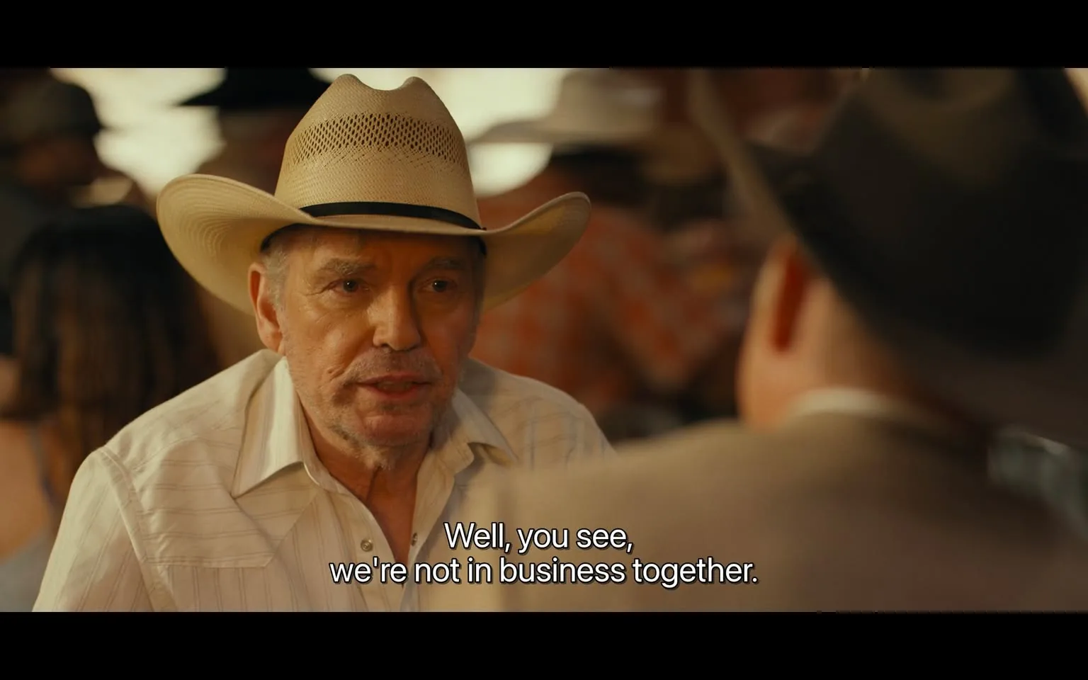 Billy Bob Thornton is prominently featured in this image, wearing a straw cowboy hat and a light-colored, button-up shirt. His expression is focused and slightly intense as he engages in conversation, suggesting a serious or important discussion. The setting appears to be indoors, likely a rustic or western-themed venue, given the other patrons in the background wearing similar cowboy hats and attire. The lighting is warm, casting a soft glow that complements the earthy tones of the setting. The background is slightly blurred, focusing attention on Thornton as the central subject. The subtitles at the bottom read, "Well, you see, we're not in business together," indicating a dialogue-driven scene, possibly from a film or television show where Thornton's character is addressing business relations. The mood is intimate yet tense, capturing a moment of negotiation or revelation. The color palette and costume design contribute to a classic Americana or Western atmosphere.
