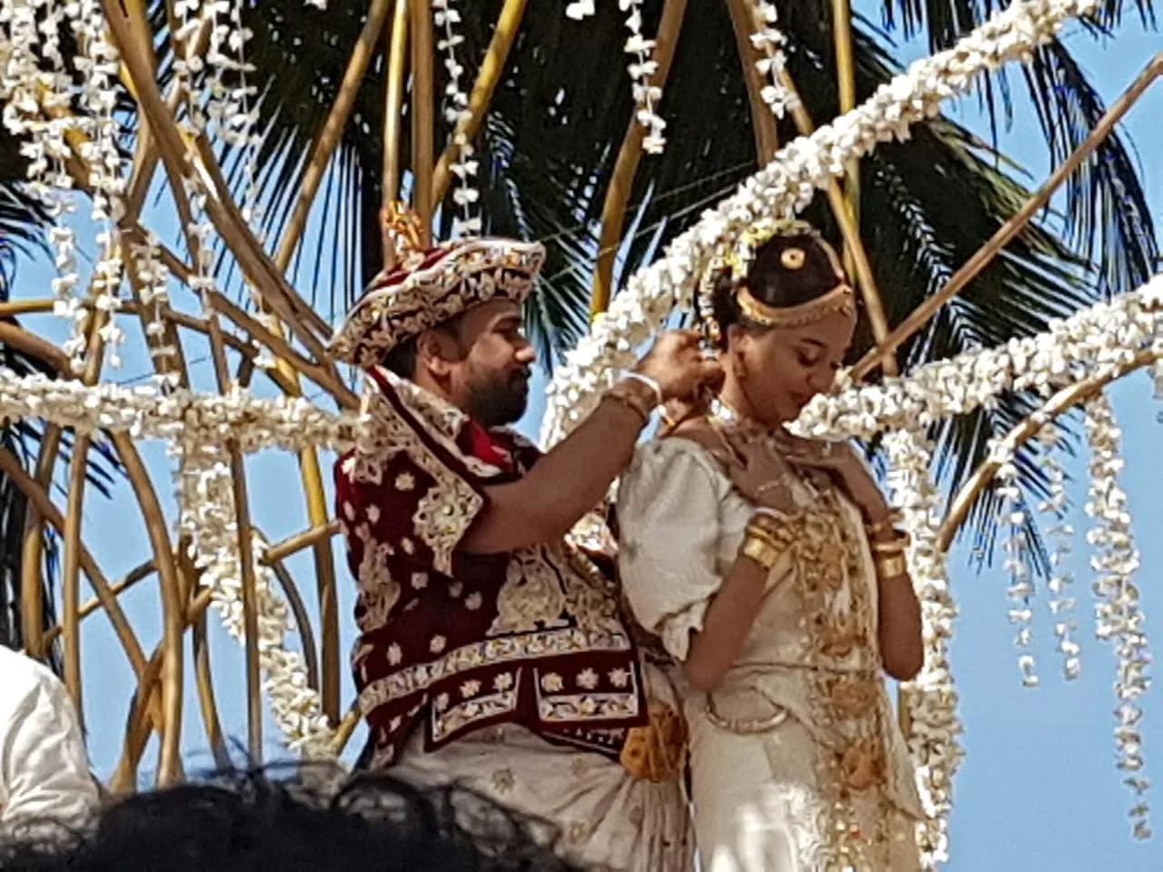 In this image, a traditional wedding ceremony is taking place outdoors under a bright blue sky. The setting is beautifully decorated with strands of white flowers hanging from a wooden structure, creating an elegant and festive atmosphere. A man, who could be identified from the hint as Raja Hettiarachchi, is engaged in the ceremonial act of placing a necklace on the bride. The groom is wearing an ornate maroon outfit with intricate gold embroidery, paired with a matching traditional hat. The bride, adorned in a resplendent white saree with gold embellishments, stands with her hands gracefully clasped near her chest, her expression serene and composed. Her jewelry is abundant and detailed, complementing her attire. Palm fronds can be seen in the background, suggesting a tropical location. The lighting is natural, highlighting the vibrant colors and fine details of their traditional clothing. The scene captures a moment of cultural significance, marked by its rich visual aesthetics and the solemnity of the occasion.

- Title: "Traditional Wedding Ceremony"