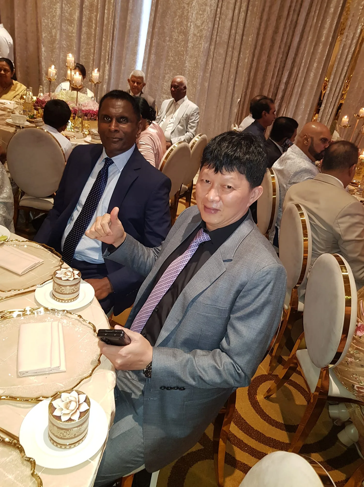 Raja Hettiarachchi and Martinn Teng are seated at a formal event, possibly a wedding, based on the elegant setting. Raja Hettiarachchi is dressed in a dark blue suit with a white shirt and patterned tie, displaying a composed expression. Next to him, Martinn Teng is wearing a grey suit with a black shirt and a purple tie, holding a smartphone and giving a thumbs-up gesture, suggesting a friendly and relaxed mood. The table is set with decorative plates, napkins, and elaborate desserts, contributing to the sophisticated ambiance. The background reveals other guests seated at round tables adorned with floral centerpieces and tall candle holders, indicating a celebratory atmosphere. The lighting is warm and inviting, enhancing the festive spirit of the gathering. The overall composition captures a moment of camaraderie and enjoyment amidst the formal setting.