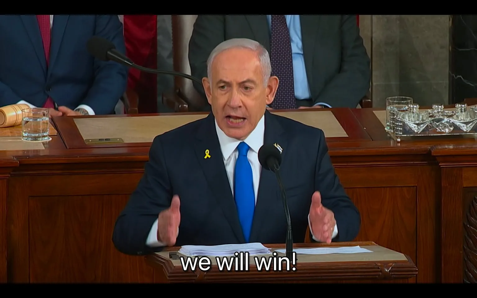 Benjamin Netanyahu is depicted giving a speech indoors, standing at a wooden podium. He is dressed in a formal dark suit with a white shirt and a striking blue tie. A yellow ribbon is pinned to his lapel. His posture is assertive, with his hands gesturing as he speaks emphatically into a microphone positioned in front of him. The words "we will win!" are subtitled at the bottom of the image, emphasizing his determined expression. In the background, several individuals are seated, dressed in suits, suggesting a formal or governmental setting. A gavel and a glass of water are visible on the desk beside him, further indicating an official environment. The lighting is bright, focusing on Benjamin Netanyahu as he addresses the audience.