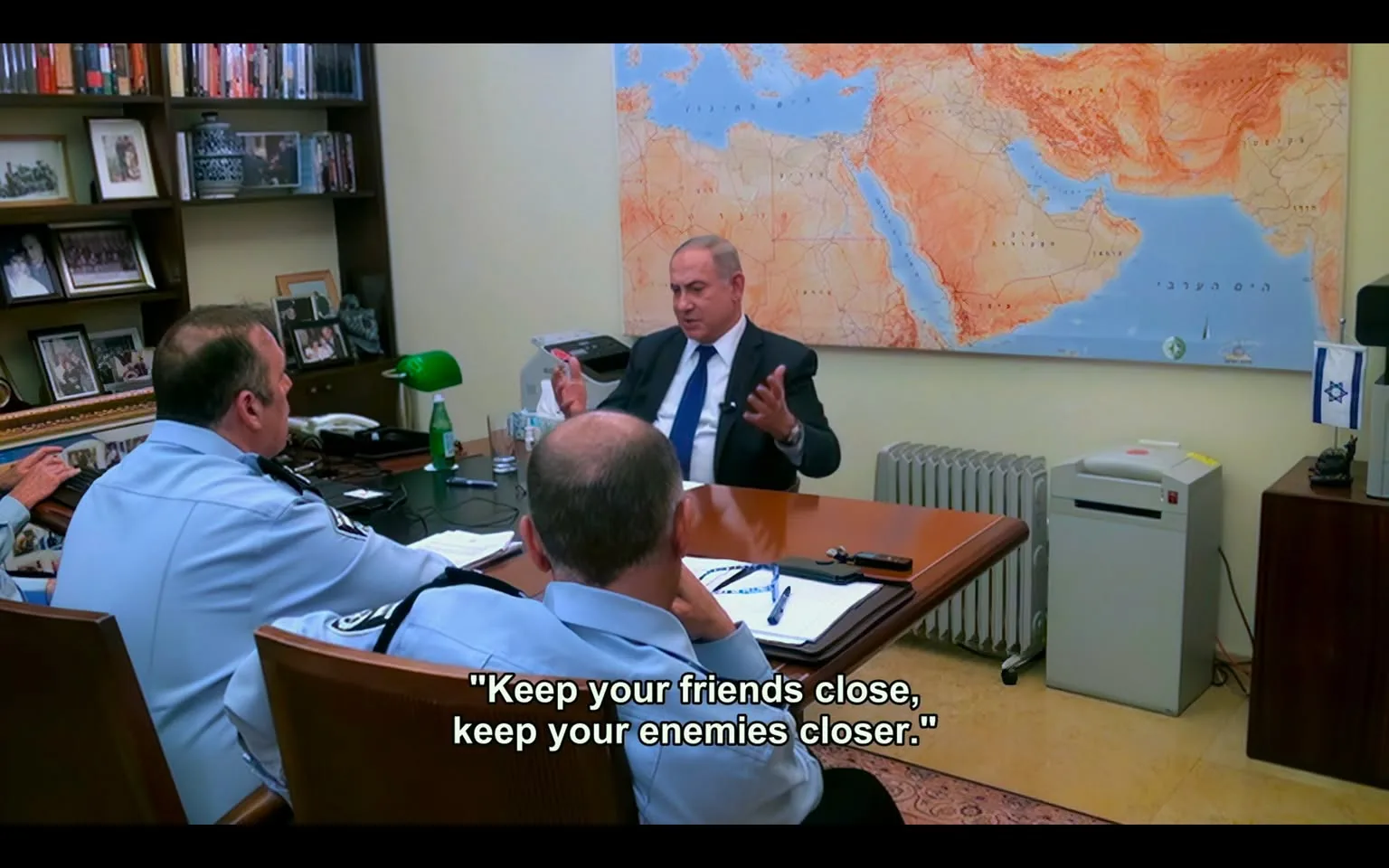 Benjamin Netanyahu is seated at a polished wooden desk in a well-organized office setting. The room appears professional, with Netanyahu gesturing expressively, possibly emphasizing a point during a discussion. Behind him is a large map of the Middle East, suggesting geopolitical discussions. On the desk are various items, including a green desk lamp, a bottle of water, and paperwork, indicating a working environment.

Two individuals in uniform, possibly officials or security personnel, are seated across from him, paying close attention. Their presence suggests a formal meeting, likely involving security or strategic discussions. Several personal photographs and books are visible on the shelves behind them, adding a personal touch to the room. An Israeli flag stands on a small table, reinforcing the national context. The ambiance is serious and focused, with subdued colors and lighting enhancing the formal atmosphere.

- Captions / subtitles: "Keep your friends close, keep your enemies closer."