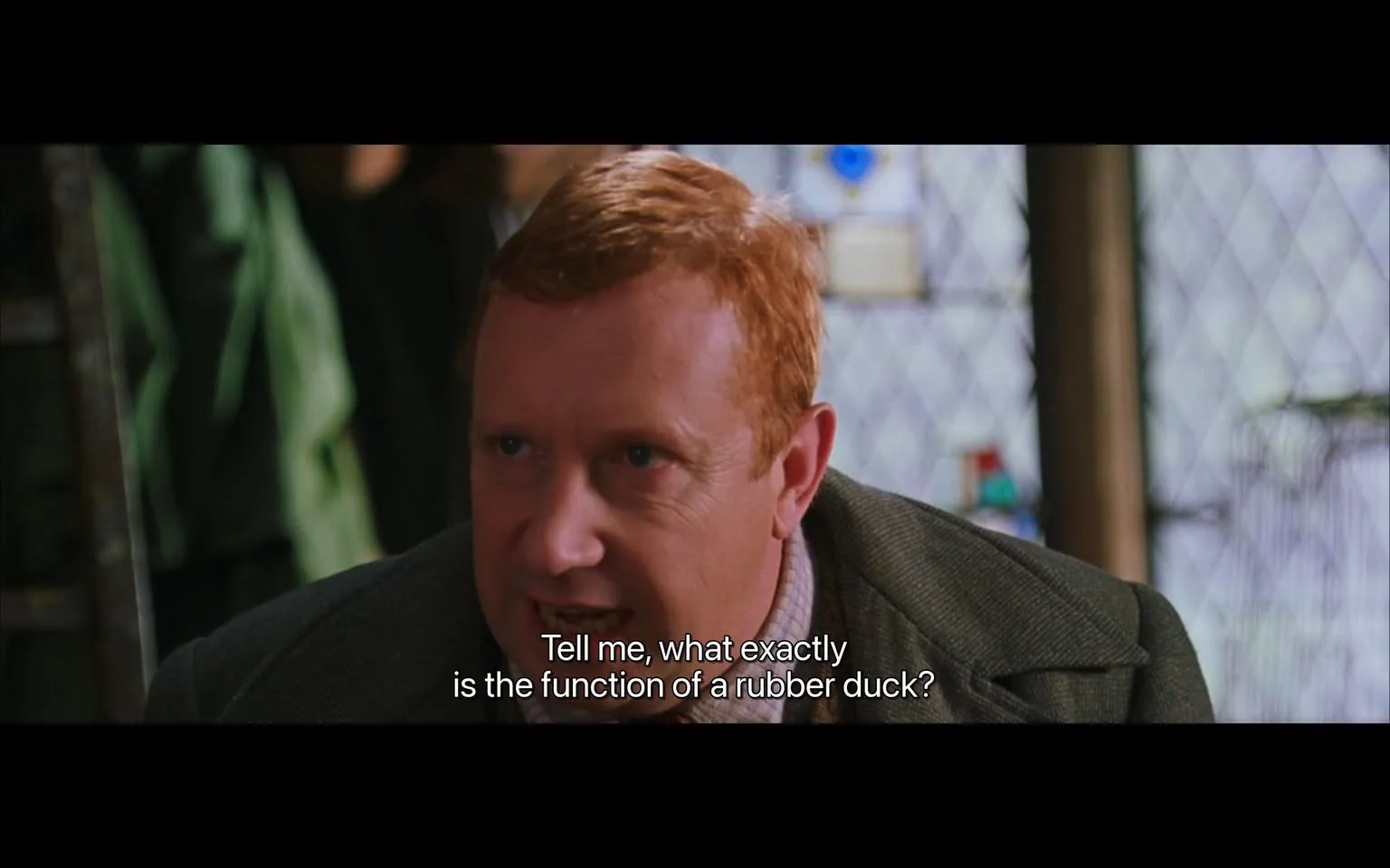 In this scene from the "Harry Potter" series, a character is shown in close-up view, engaging in a conversation indoors. The character has a focused expression and is leaning slightly forward, suggesting curiosity or puzzlement. He has reddish hair and is dressed in a brown tweed suit, adding to the sense of a casual or academic setting. The backdrop features a diamond-patterned window and some indistinct objects, contributing to a cozy, somewhat cluttered atmosphere typical of wizarding environments. The lighting is soft, enhancing the intimate mood of the exchange. The caption "Tell me, what exactly is the function of a rubber duck?" adds a humorous element, as it implies a lack of understanding of everyday Muggle items. This reflects the whimsical and often comedic contrasts between the magical and non-magical worlds in the series.