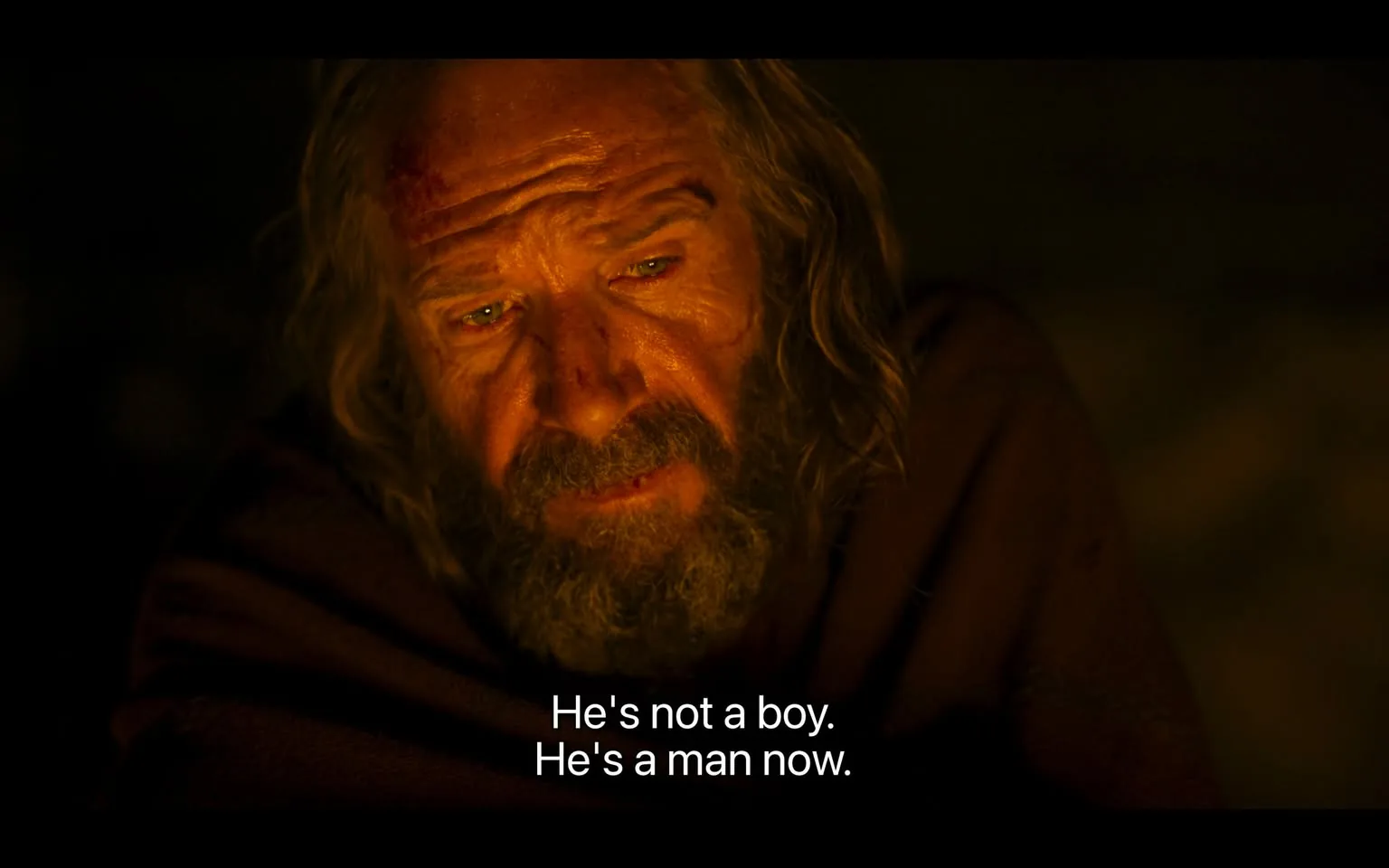 1) Rich description: The image features Ralph Fiennes in a scene from the film "The Return" (2024). He appears to be in a dimly lit setting, possibly illuminated by a fire or candle, as warm tones wash over his face, emphasizing the lines and texture of his skin. His expression is somber and reflective, suggesting a moment of introspection or seriousness. Fiennes has long hair and a beard, adding to the rugged, weathered appearance of his character. The background is dark and indistinct, focusing attention on his face and the emotions being conveyed. He is draped in a cloak or heavy garment, enhancing a sense of historical or timeless context. The subtitle reads, "He's not a boy. He's a man now," indicating a pivotal moment in the dialogue. This suggests themes of growth, change, or acceptance within the narrative.

2) On-screen text block:
- Captions / subtitles: "He's not a boy. He's a man now."