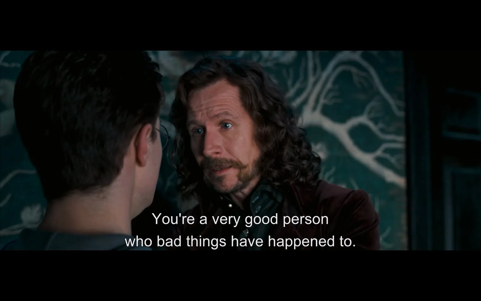 Harry Potter is depicted in a dramatic indoor scene with Sirius Black. Sirius Black, characterized by his long, wavy hair and mustache, is speaking earnestly to Harry Potter, who is seen from behind. The expression on Sirius Black's face is sincere and sympathetic, suggesting a moment of emotional connection and support. The setting features a backdrop with intricate patterns resembling tree branches, which adds a mystical element to the scene. The lighting is dim, emphasizing the serious tone of the conversation. The attire of the characters is suited to a wizarding world, with Sirius wearing a dark, formal jacket. The subtitles on screen read, "You're a very good person who bad things have happened to," indicating a moment of reassurance and empathy. This scene captures a pivotal interaction between the two characters, highlighting themes of friendship and resilience.

- Captions / subtitles: "You're a very good person who bad things have happened to."