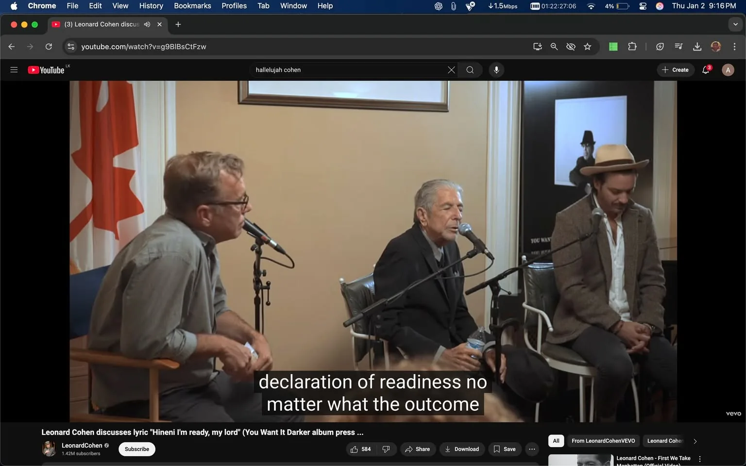 In a discussion setting, three individuals are seated with microphones in front of them. The main subject appears to be an older man with gray hair, dressed in a black suit, who is speaking into a microphone. He holds a water bottle in one hand. To his left, a man in a gray shirt and glasses listens attentively, while to his right, another man in a brown hat and a light-colored blazer appears to be engaged in the conversation. The background features a Canadian flag and a framed picture on the wall. Subtitles indicate that the discussion revolves around a lyric from a song, emphasizing themes of readiness and acceptance. The atmosphere is informal, likely part of a press event or interview.
