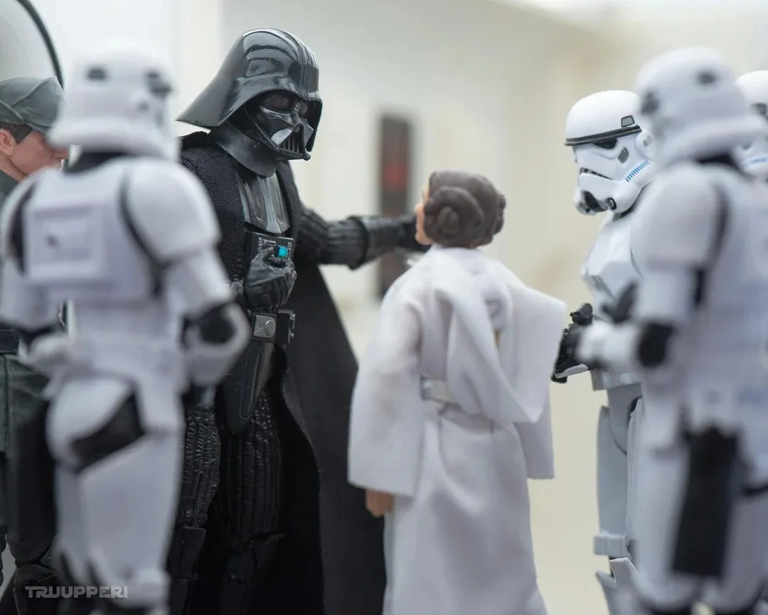 In the scene, a group of action figures is depicted in a tense moment. The central figure, dressed in a black cape and helmet, is likely a representation of a well-known character from a sci-fi franchise, gesturing towards a figure in a white robe, who appears to be in a defensive stance. Surrounding them are several figures in white armor, resembling soldiers, standing in a semi-circle. The setting appears to be indoors, with a neutral background that suggests a futuristic environment. The figures are detailed, showcasing various textures and colors, contributing to the dramatic atmosphere of the scene. The presence of weapons is implied, enhancing the sense of conflict.