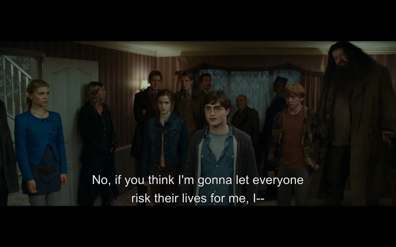 In a dimly lit room with striped wallpaper, a group of individuals stands in a tense formation, facing one central figure. The main subject, a young adult male with glasses and a casual outfit, appears to be expressing concern or frustration, as indicated by his body language and facial expression. Surrounding him are several others, including a young woman in a denim jacket, a man with a beard, and others dressed in casual clothing. The atmosphere suggests a serious discussion or conflict, with the characters displaying a range of emotions from worry to determination. The setting is indoors, with a staircase visible in the background and soft lighting creating a somber mood.