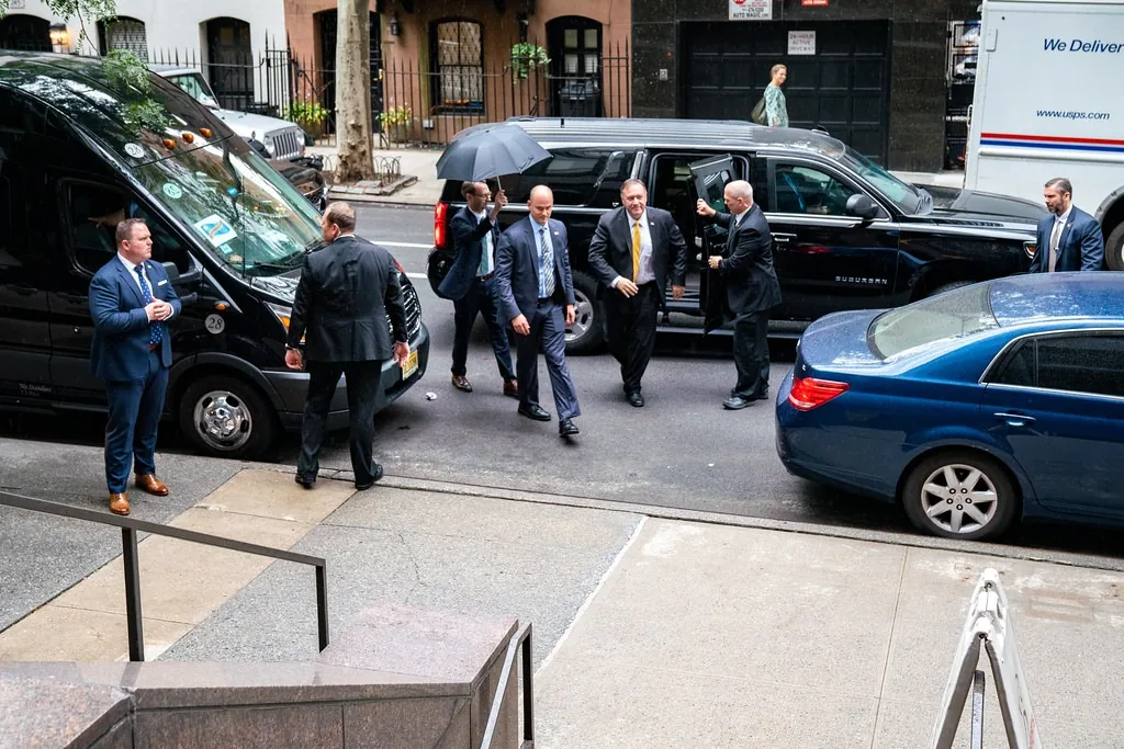 A group of men in suits is seen exiting a black vehicle, likely a security detail, in an urban setting. The men appear to be in their 30s to 50s, dressed in formal attire, with some wearing ties. One man stands by a black van, while others are engaged in conversation or preparing to enter another vehicle. The background features a USPS delivery truck, indicating a city environment. The scene suggests a high-profile event or individual, given the presence of security personnel. The weather appears overcast, and the street is lined with buildings typical of an urban area.