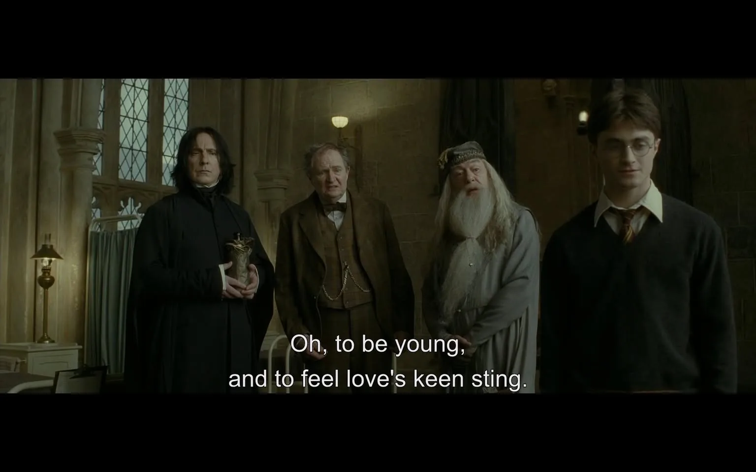 In a dimly lit indoor setting, several characters are present, engaged in conversation. The scene captures a moment of reflection, with one character expressing a sentiment about youth and love. The characters include an adult male with long dark hair and a serious expression, wearing a black robe, holding a decorative object. Another adult male, dressed in a brown suit, appears contemplative. A third character, an older man with a long white beard, is dressed in gray robes, while a younger male, wearing glasses and a dark sweater, stands slightly apart, looking thoughtful. The background features stone walls and large windows, suggesting a historical or academic environment. The dialogue displayed on the screen reads, "Oh, to be young, and to feel love's keen sting."