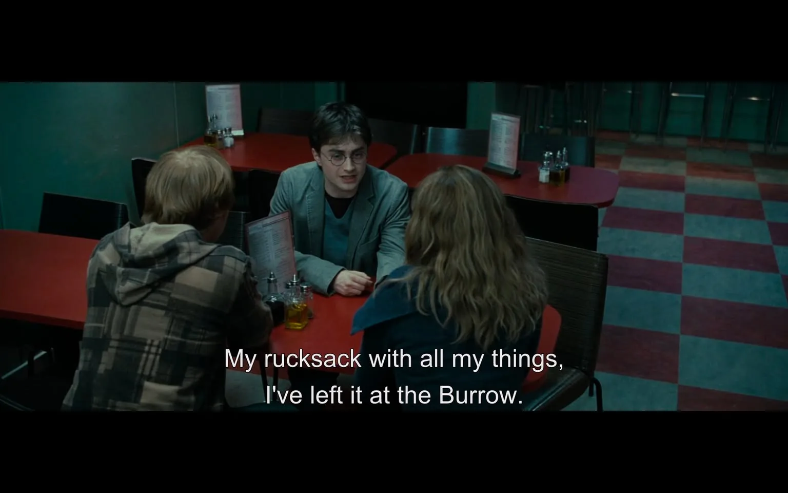 In a casual dining setting, three individuals are seated at a red table. The focus is on one person, who is wearing glasses and a gray jacket, engaged in conversation. He appears to be explaining something important, mentioning a rucksack left at "the Burrow." The other two individuals, one with short hair in a plaid hoodie and the other with long, wavy hair, listen attentively. The background features menus on the table and a tiled floor with a checkerboard pattern. The atmosphere seems informal, likely during a meal or a break.